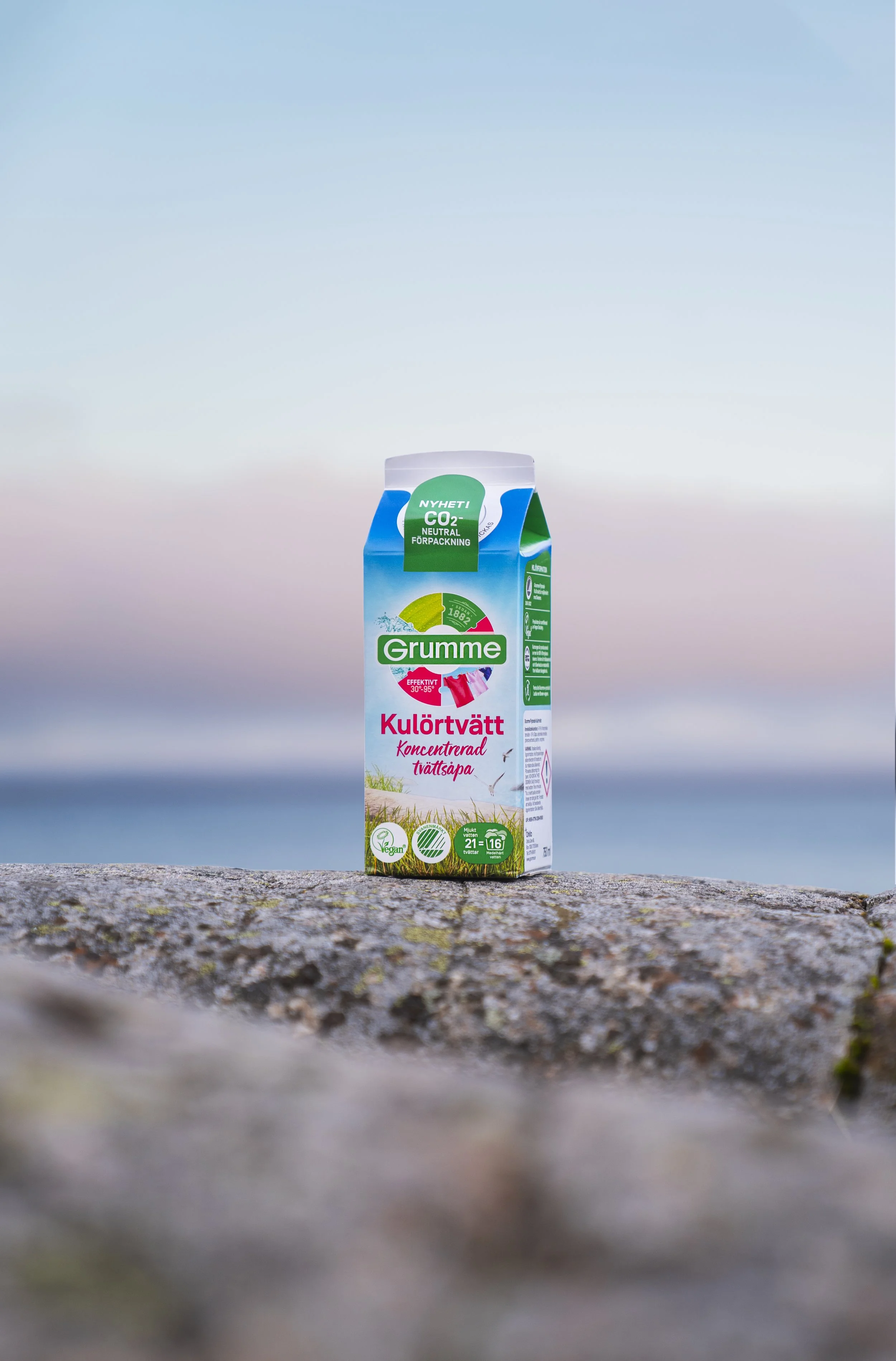 A carton of Grumme concentrated laundry detergent with a scenic beach background and a blurred rocky foreground.