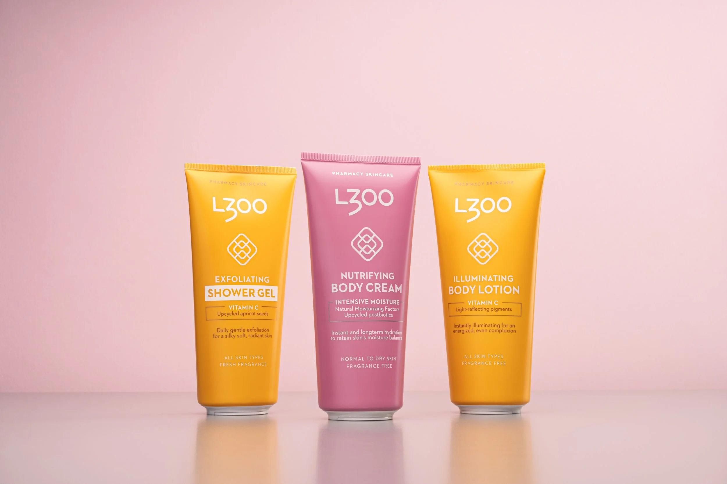 Three skincare product bottles on a pink background: a yellow exfoliating shower gel, a pink nourishing body cream, and a yellow illuminating body lotion.