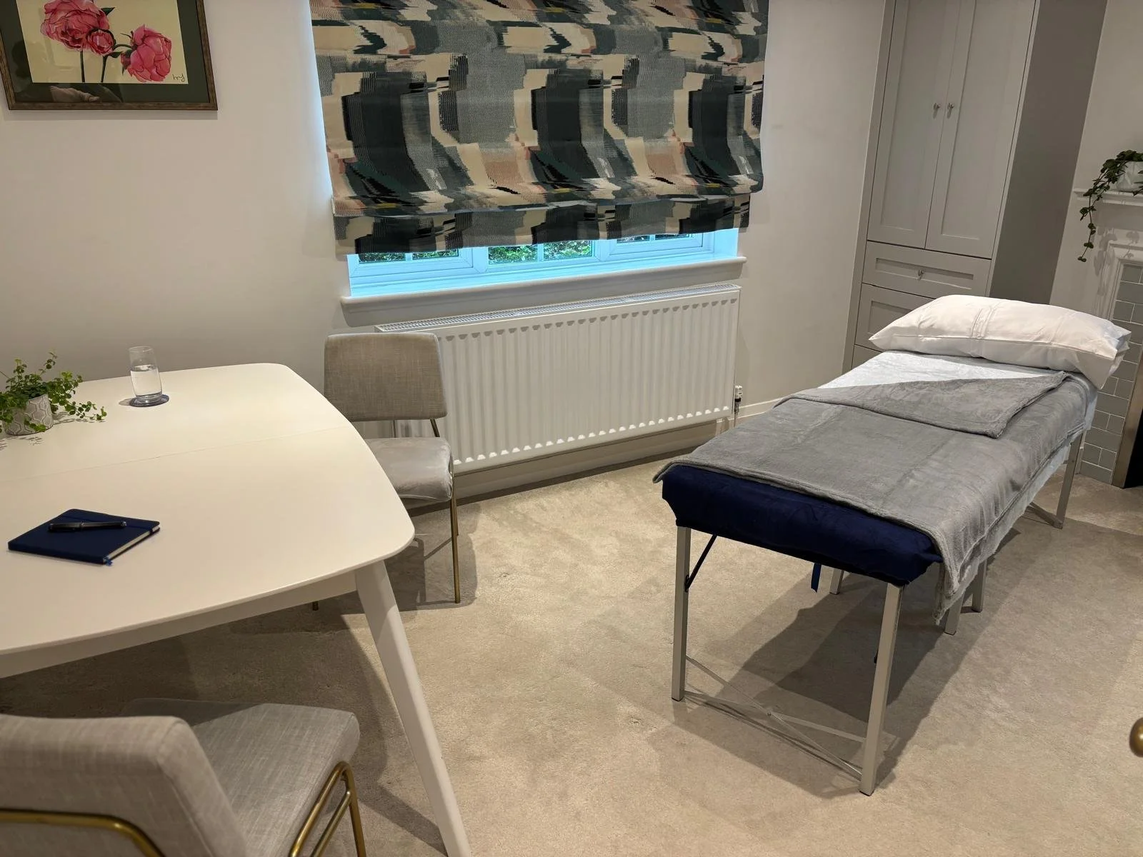 A room with a therapy or examination table covered with a gray sheet and pillow, a round white table with a blue notebook and pen, two beige chairs, a window with a patterned curtain, a white radiator below the window, and a white storage cabinet in 
