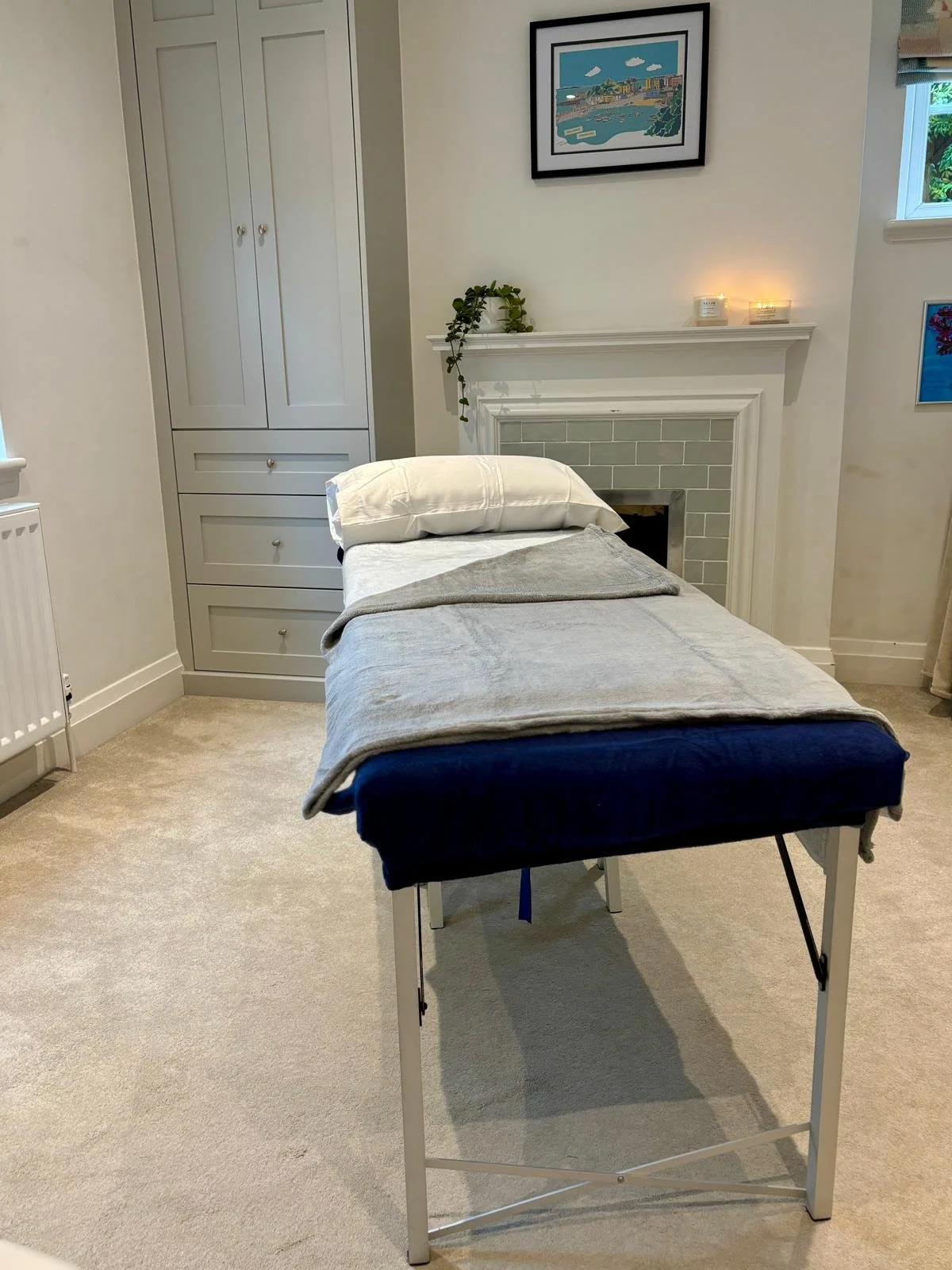 A massage or treatment table with a pillow and blankets in a cozy room with light-colored walls, a small fireplace, framed artwork on the walls, candles on the mantel, a window with blinds, and a tall closet or cabinet.
