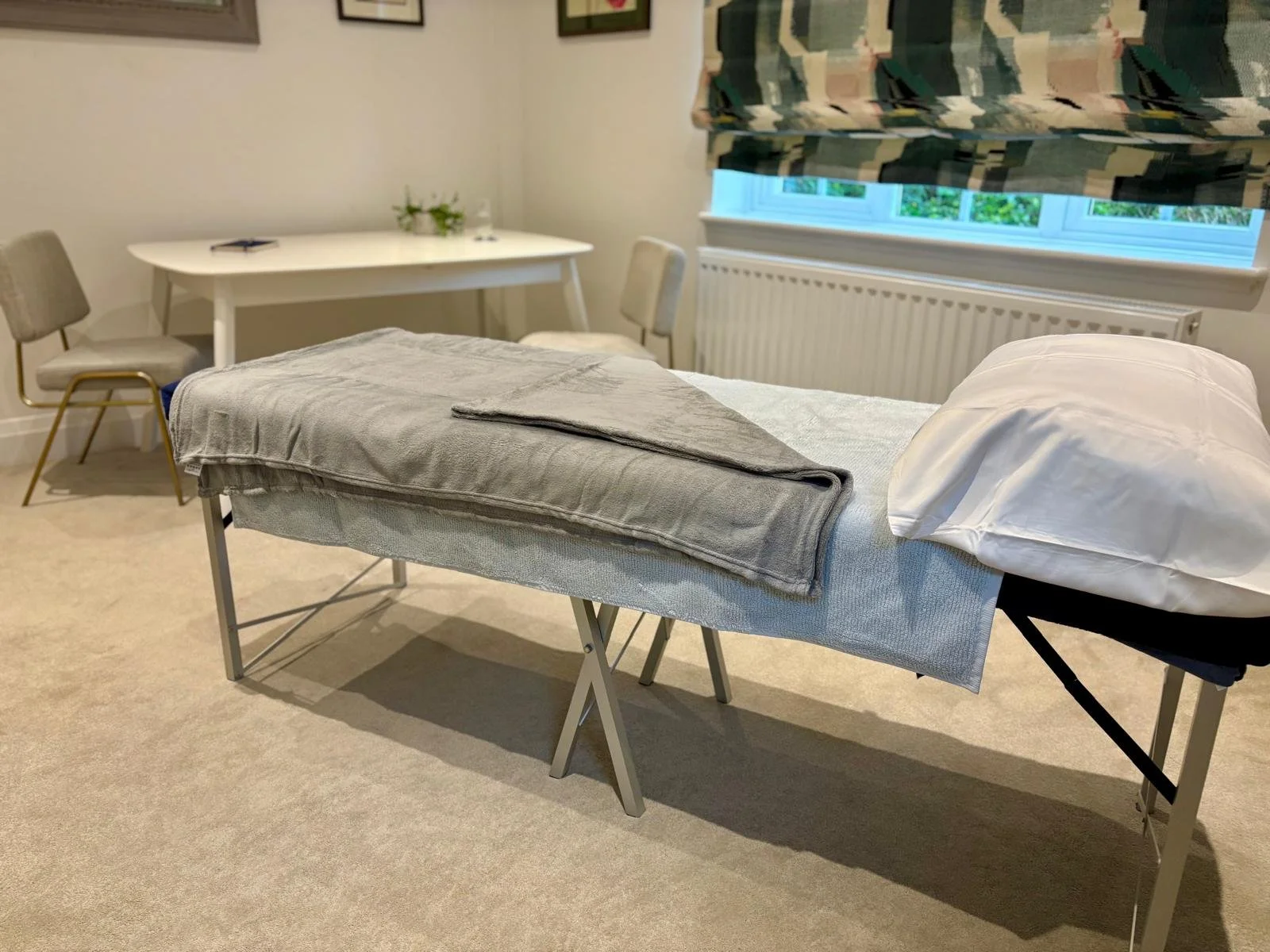 A massage table with a folded gray towel and white pillow in a therapy room. There is a white table with chairs and a small vase with flowers in the background, and a window with a patterned valance.
