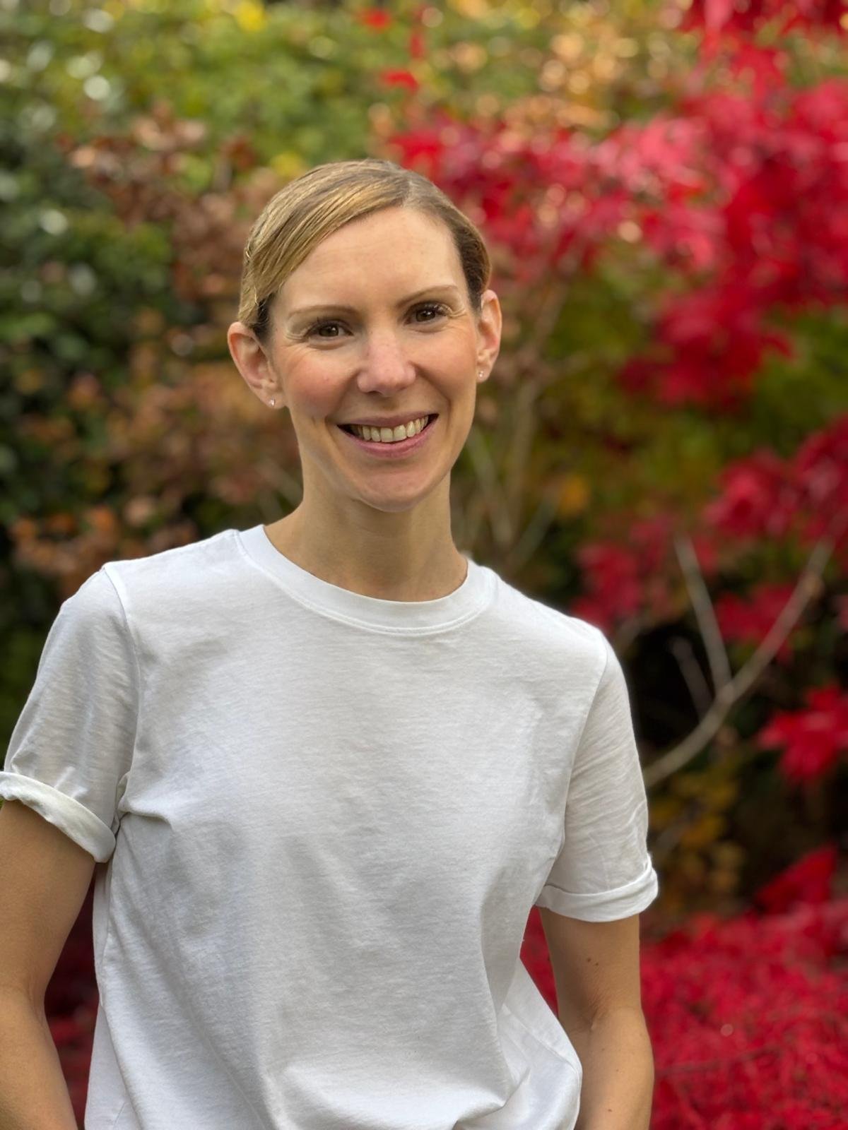 Smiling woman with short hair in a white t-shirt outdoors with colorful red and green foliage in the background.