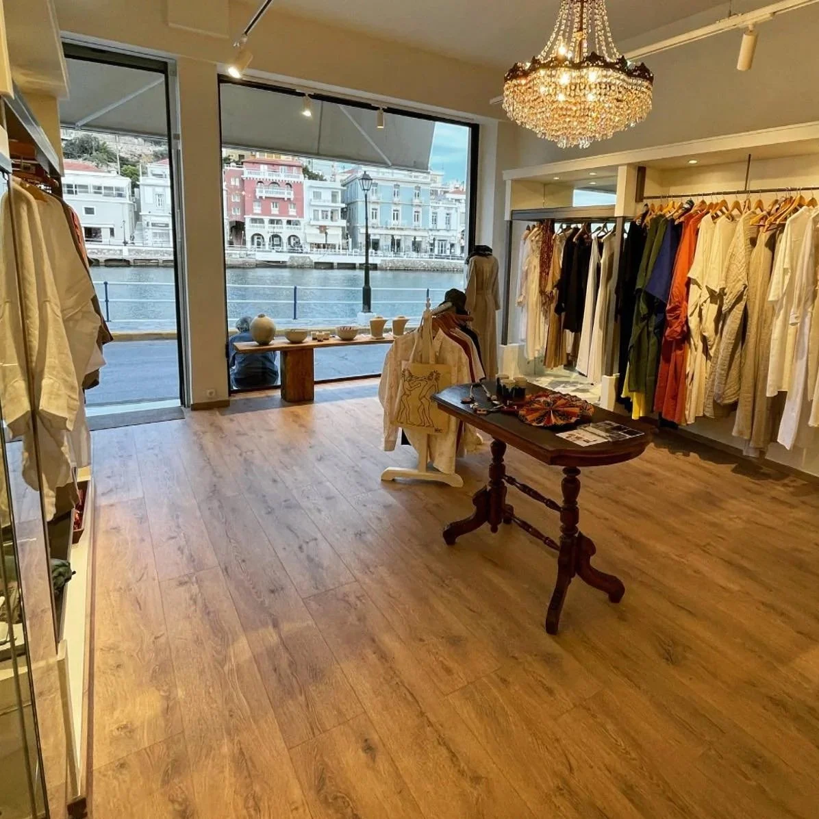 Vendemma Concept store - A clothing boutique with large windows showing a river and colorful buildings across the water. The store has wooden flooring, racks of hanging clothes, a table with accessories, and a chandelier overhead.