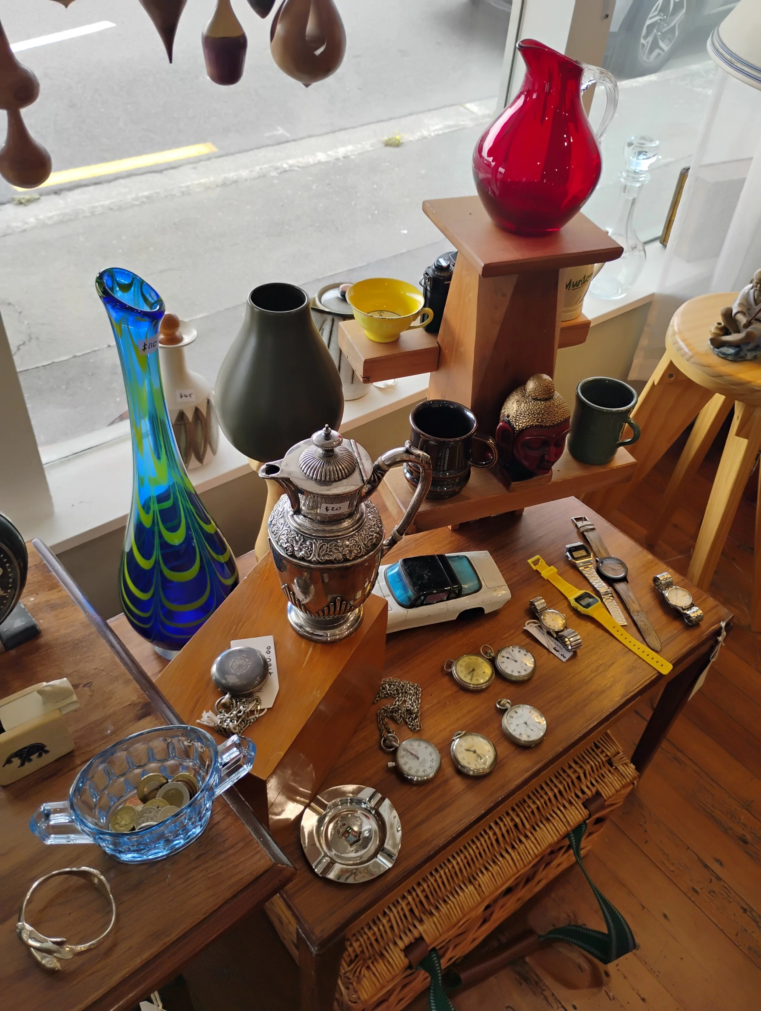 Display of various decorative glassware, ceramics, watches, and small collectibles in a shop window, with a street visible outside. chch antique shop