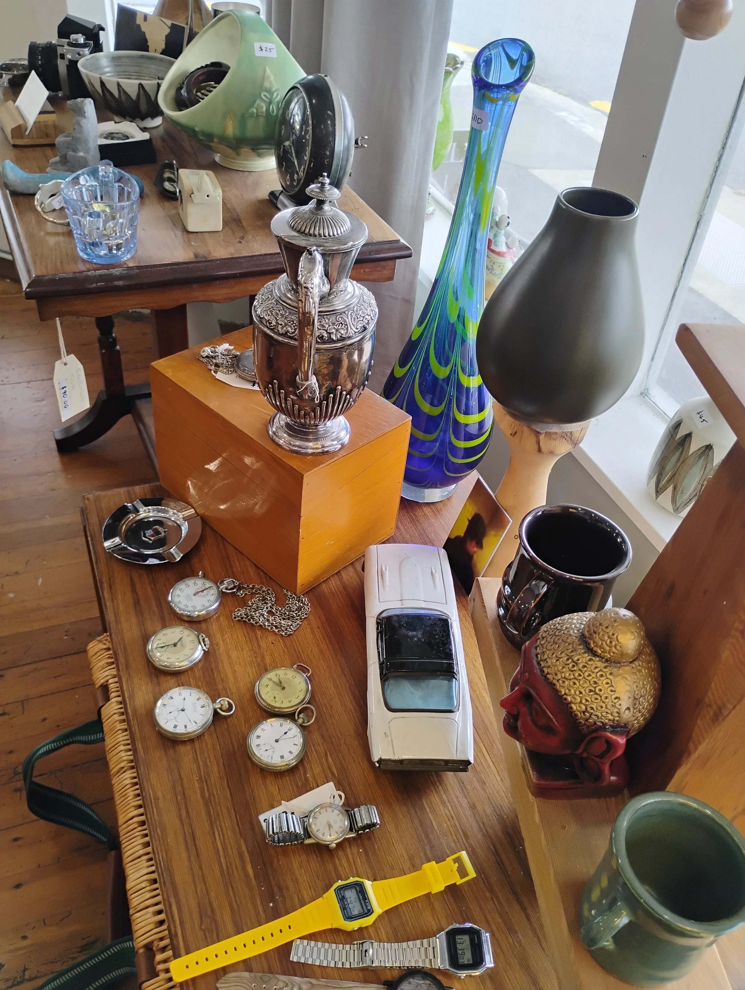 A wooden table displaying various antique and vintage items, including pocket watches, a model car, a ceramic head sculpture, a yellow digital watch, cups, mugs, vases, a clock, and other decorative objects.
