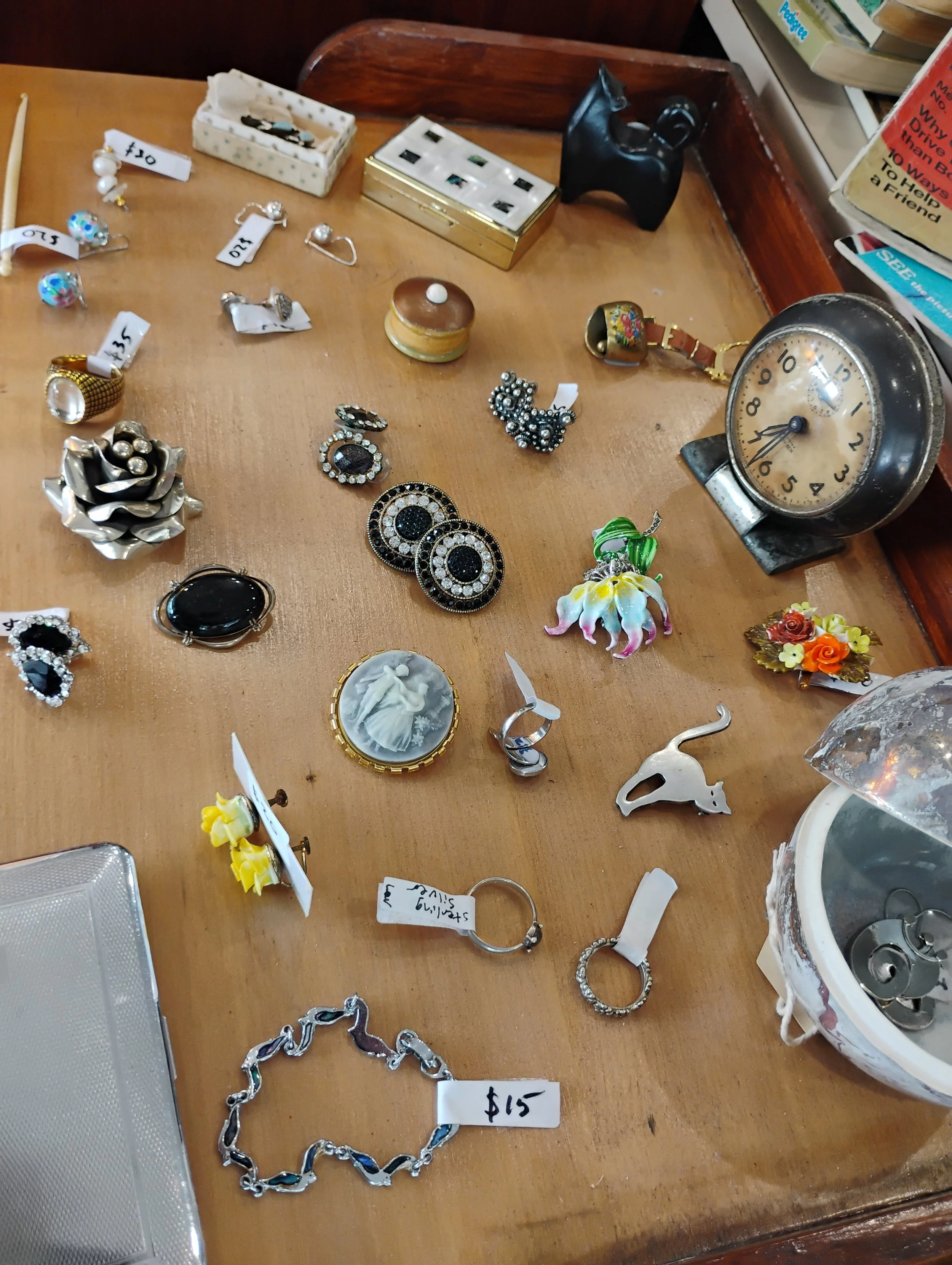 A wooden table displaying various vintage jewelry, rings, brooches, and a clock, with small price tags attached. In the background, there are books and a small black figurine.