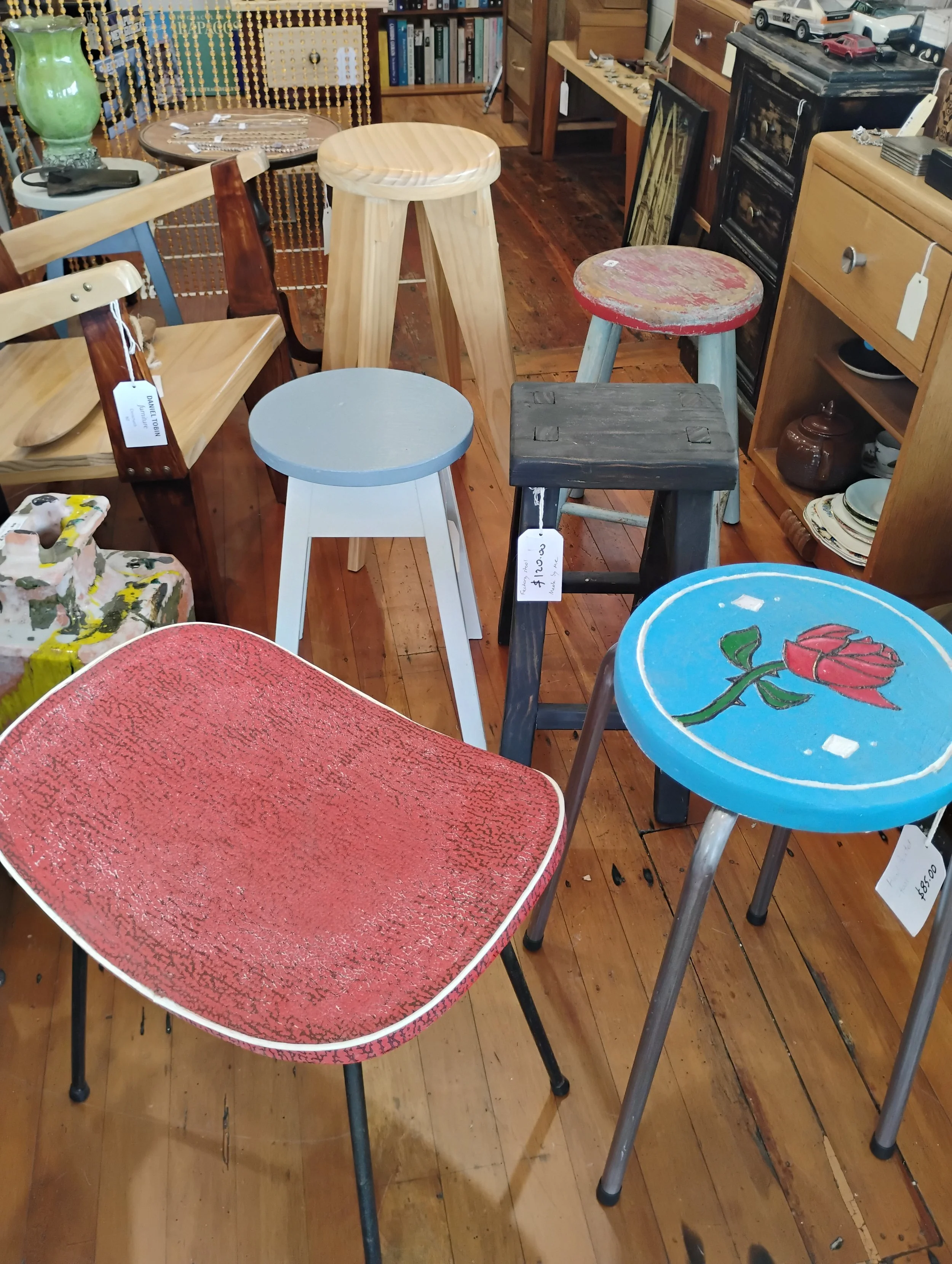 Various vintage and decorative stools and chairs on a hardwood floor in a thrift store or antique shop. Items include a red textured seat, a blue round seat with a rose painting, a black wooden stool, and a gray stool, with books and furniture in the