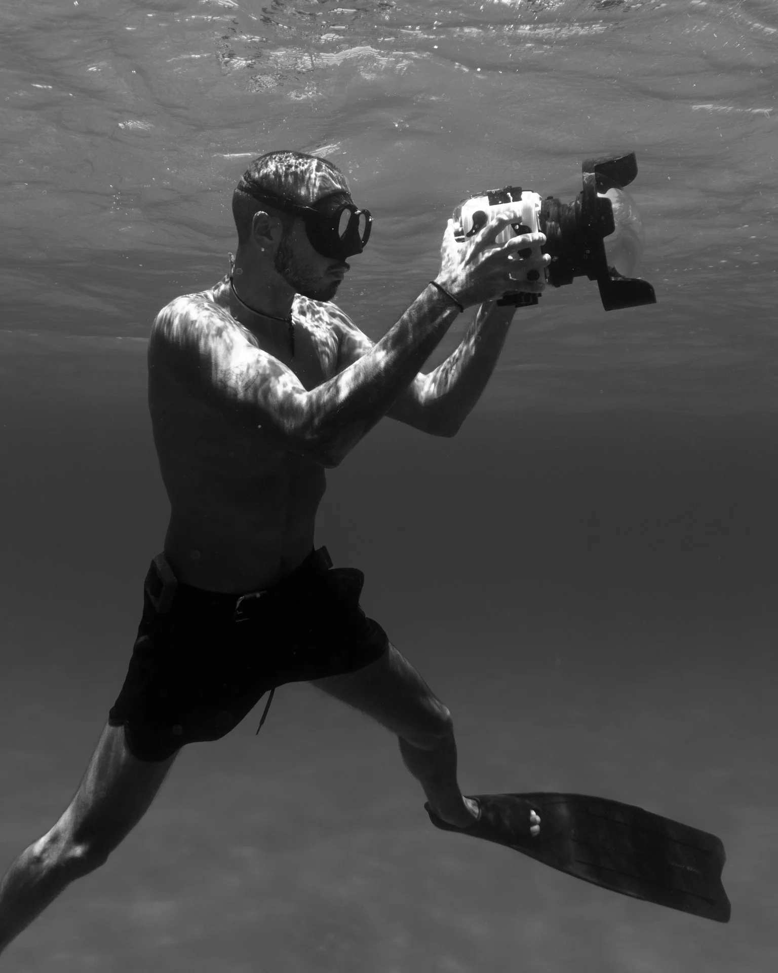 A man underwater wearing goggles and shorts, holding a camera with a waterproof housing and a wide-angle lens, taking a photograph.