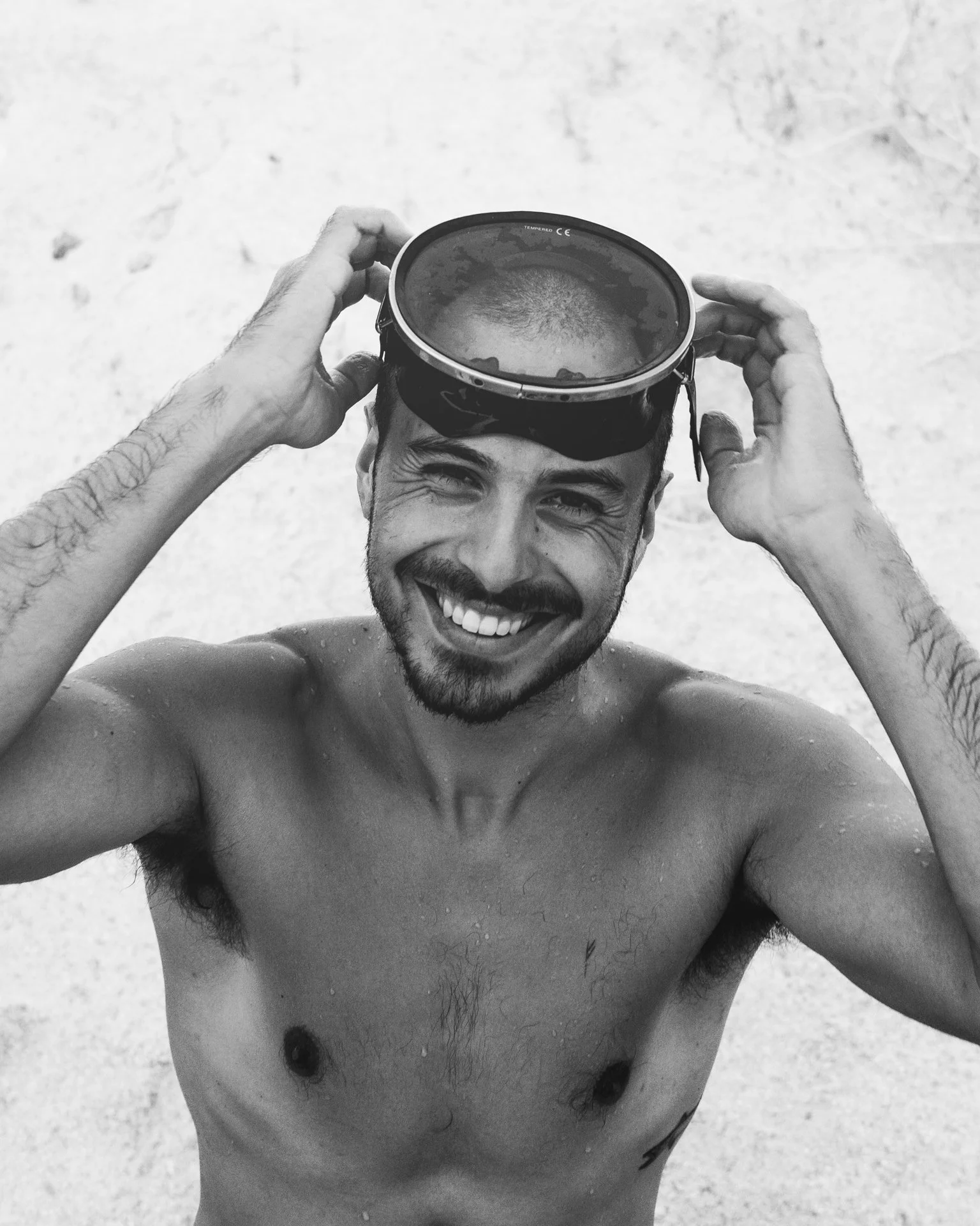 A smiling, shirtless man with a beard and tattoos on his arms, holding snorkeling goggles on his head on a beach in black and white.