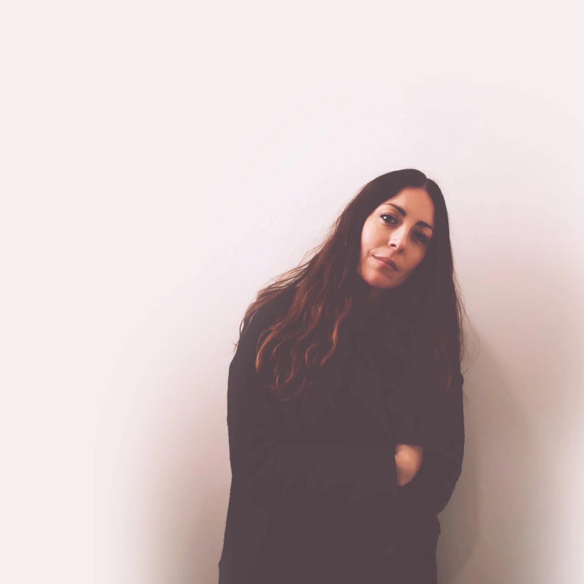 A woman with long wavy hair, wearing a black top, standing with arms crossed against a plain white wall.