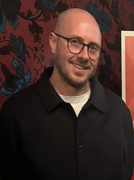 A smiling man with glasses, a beard, and a shaved head, wearing a black shirt, standing in front of a dark red and black patterned background.