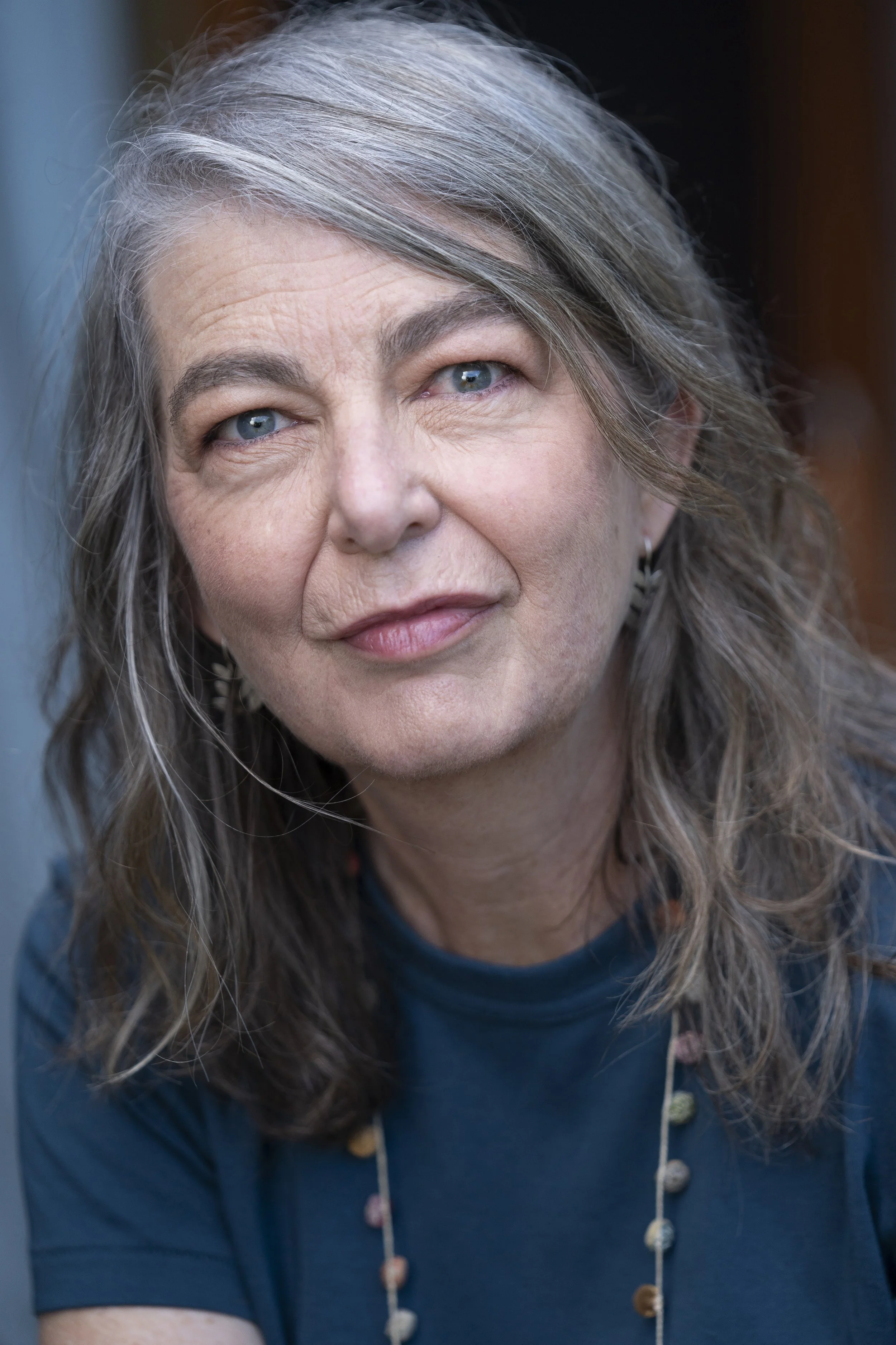 Close-up of a mature woman with gray, wavy hair and blue eyes, wearing a dark blue top and beaded necklace, looking directly at the camera.