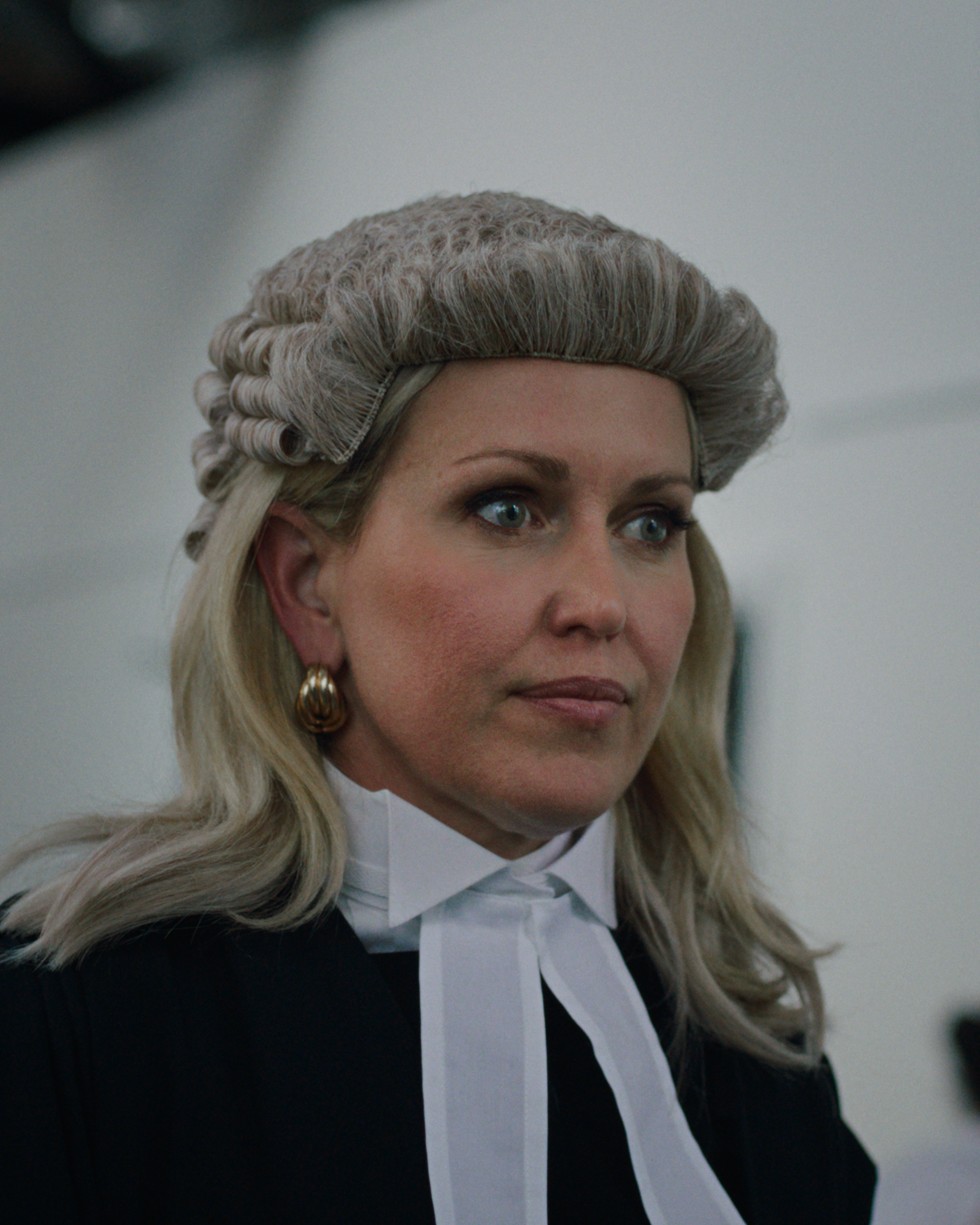 Jennifer Robinson dressed as a judge with a gray wig, white collar, black robe, and gold earrings, looking serious in a courtroom