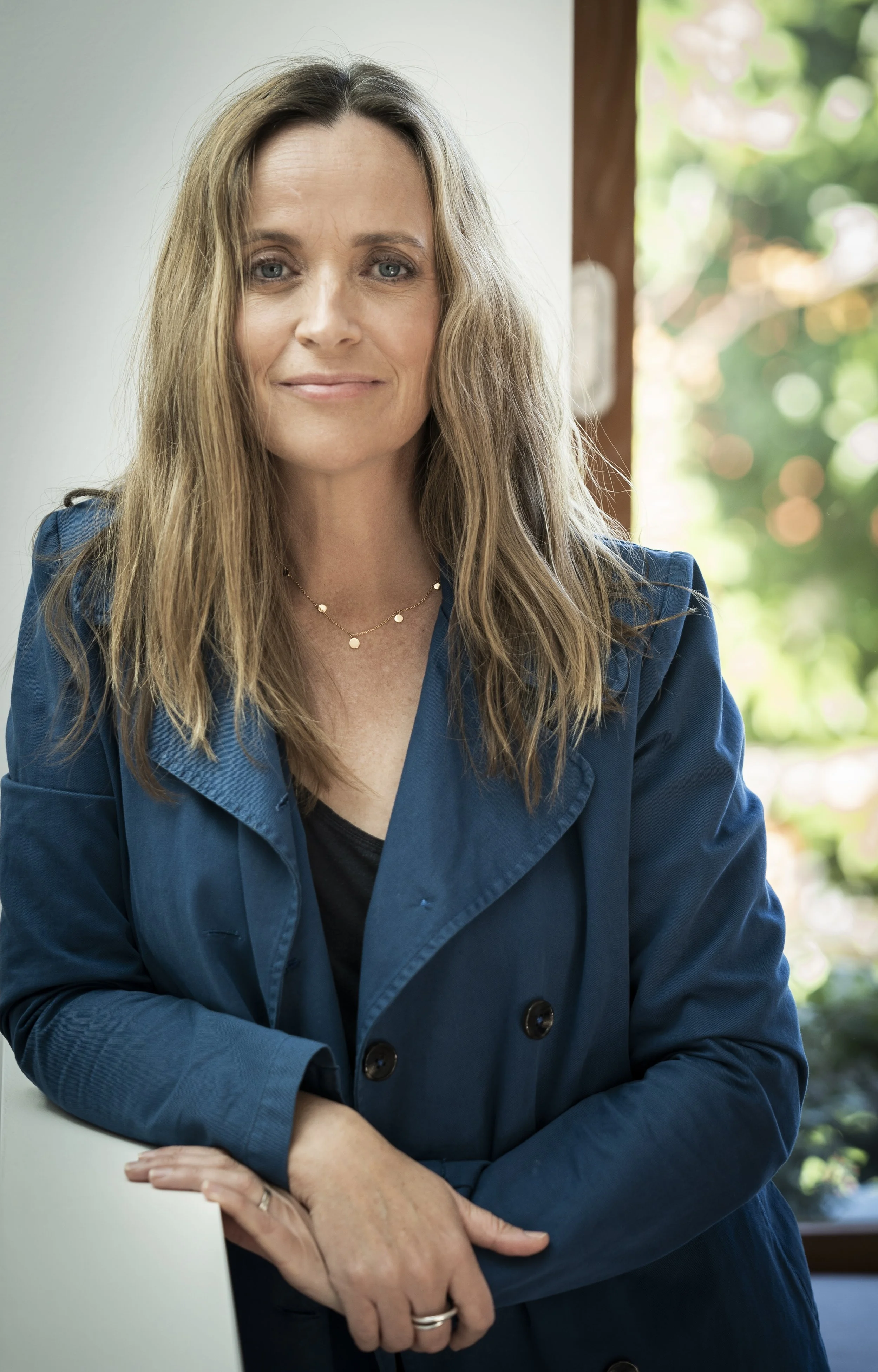 A woman with long, wavy blond hair wearing a navy blue blazer and a black top, standing indoors near a window with greenery outside, smiling gently.