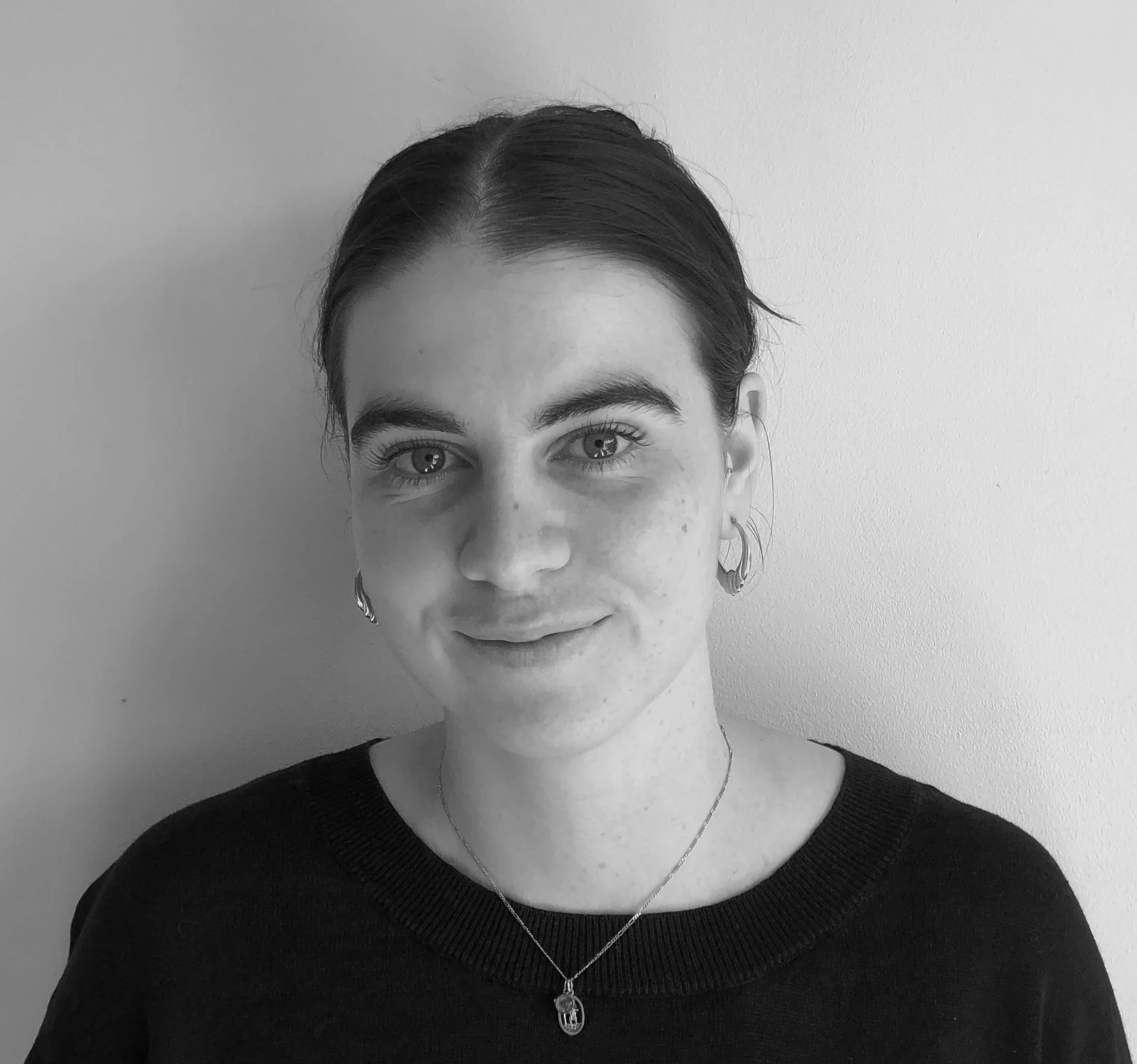 Black and white portrait of a young woman with dark hair pulled back, wearing earrings, a necklace, and a dark top, smiling gently at the camera against a plain wall background.