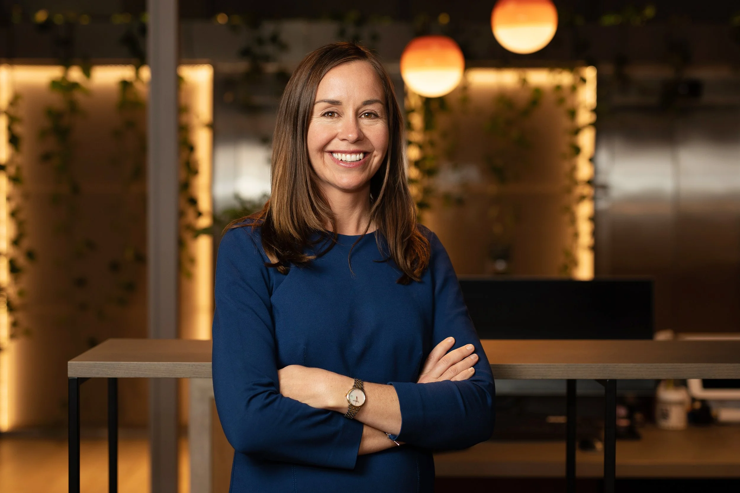 A woman with brown hair, smiling, wearing a blue long-sleeve top and a watch, standing with her arms crossed in a modern office space with warm lighting and decorative plants in the background.