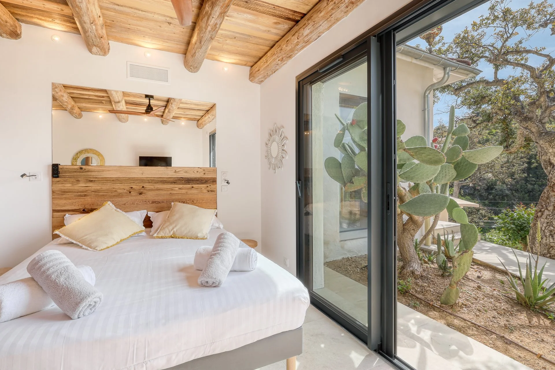 Chambre moderne avec lit blanc, oreillers crème, serviettes roulées, grande fenêtre en verre, vue sur un arbre cactus et paysage extérieur