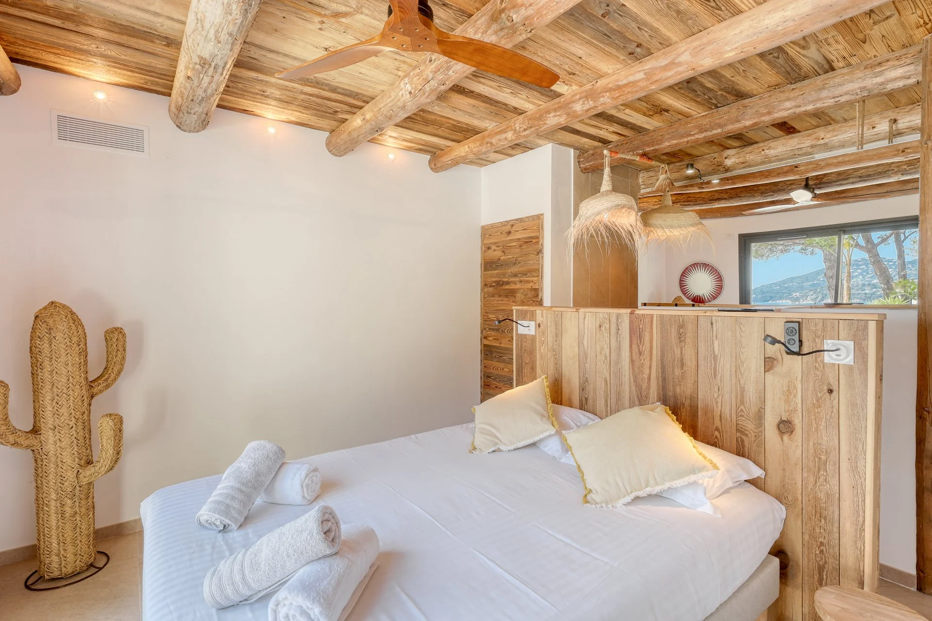 Chambre avec lit, oreillers, serviettes, décor en bois, plafond en poutres en bois, lampe en rotin, fenêtre montrant un paysage avec des arbres et des montagnes.