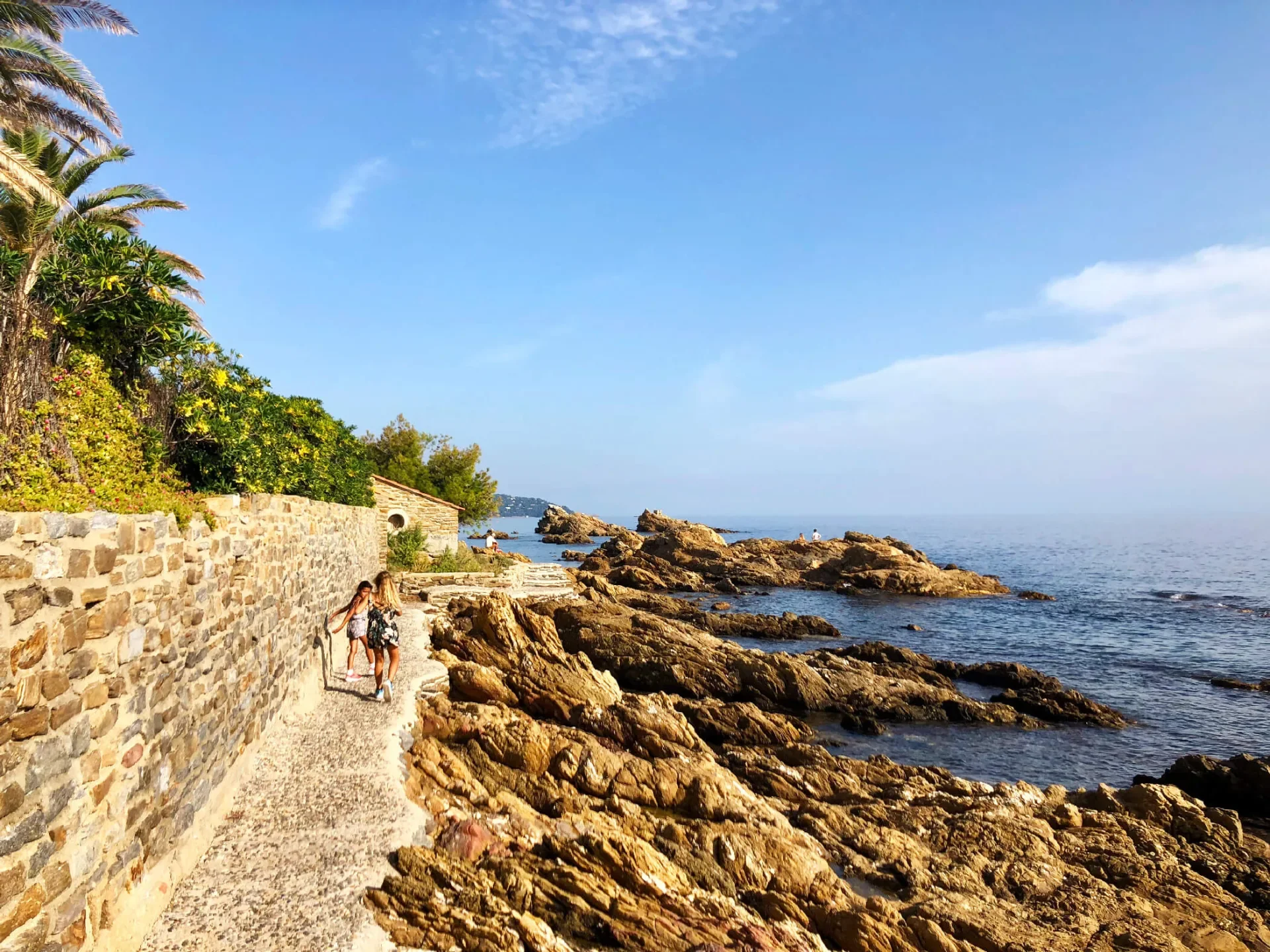 Deux femmes marche sur un sentier côtier en pierre, bordé de murs en pierre et de végétation luxuriante, face à la mer avec des rochers et un ciel partiellement nuageux.