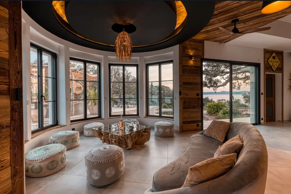 Salon moderne avec grand canapé, ottomans, table en bois, fenêtres panoramiques offrant vue sur la nature et l'eau, décoration en bois et têtes de lampes suspendues en raphia.