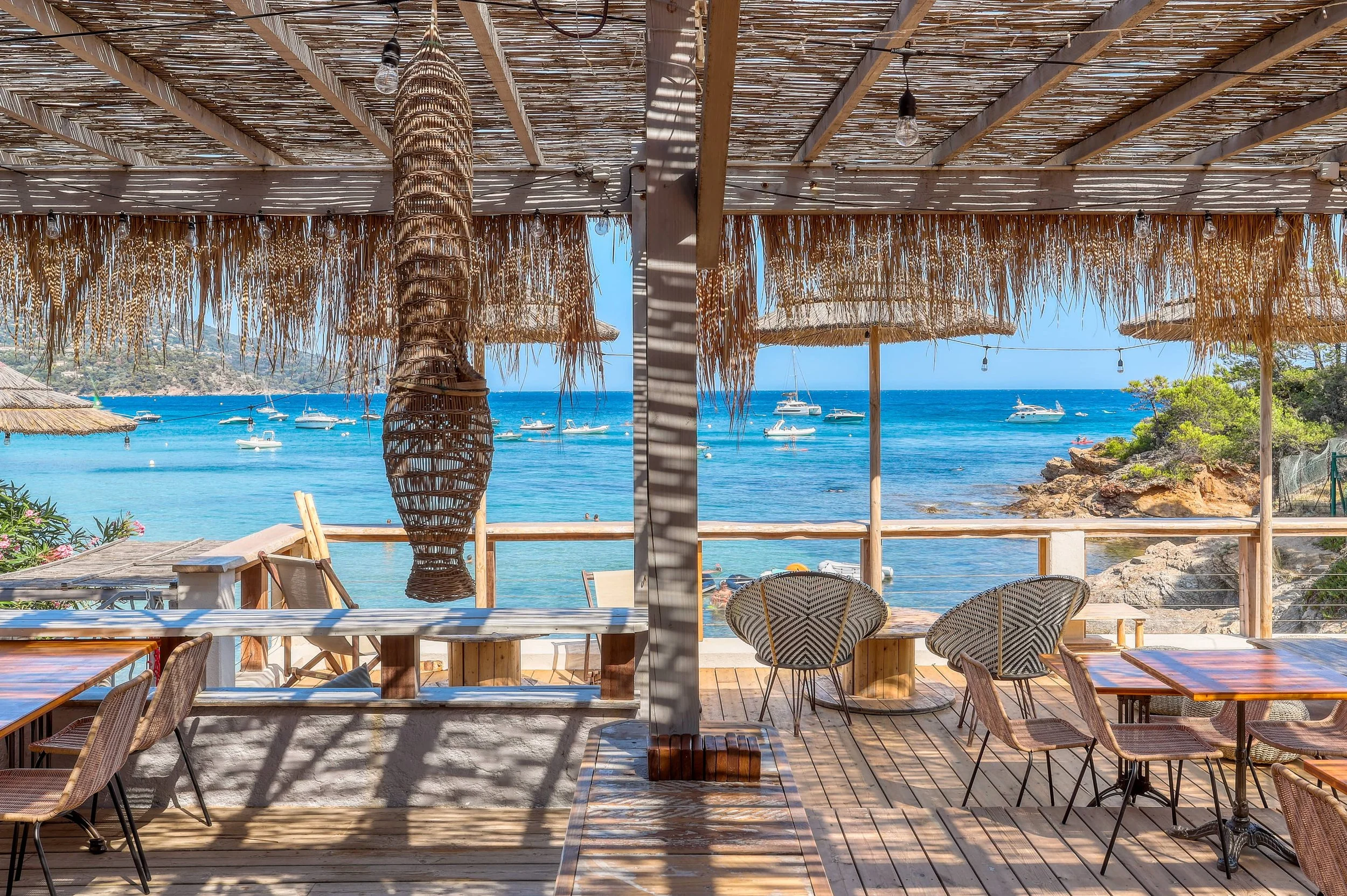 Une terrasse en bois avec des chaises et tables face à la mer, avec des parasols en paille et une vue sur des bateaux dans l'eau.