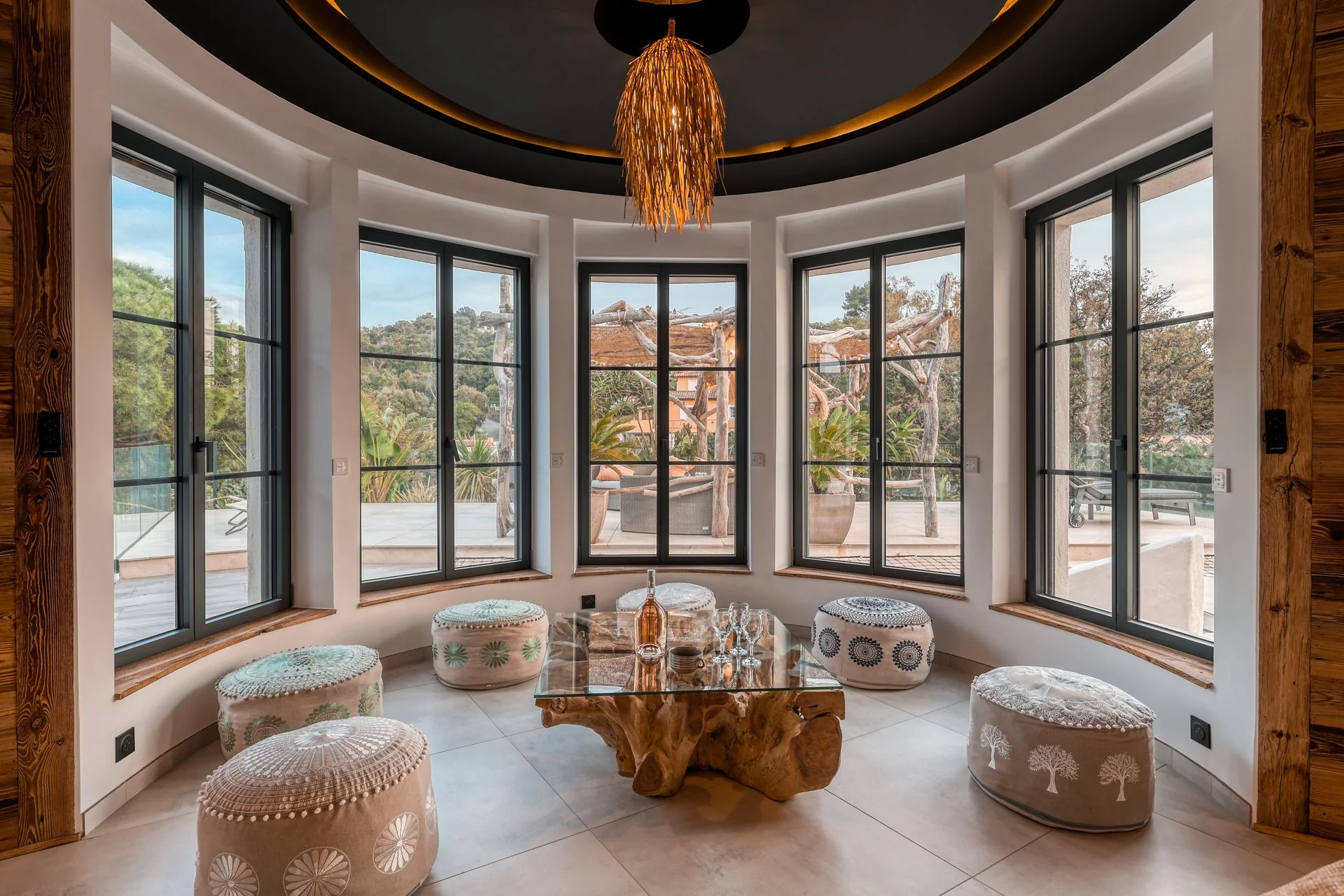 Salle de séjour avec une grande baie vitrée, plafond noir avec un lustre en bois, table en bois massif, poufs décoratifs autour, vue sur un patio avec végétation extérieure.