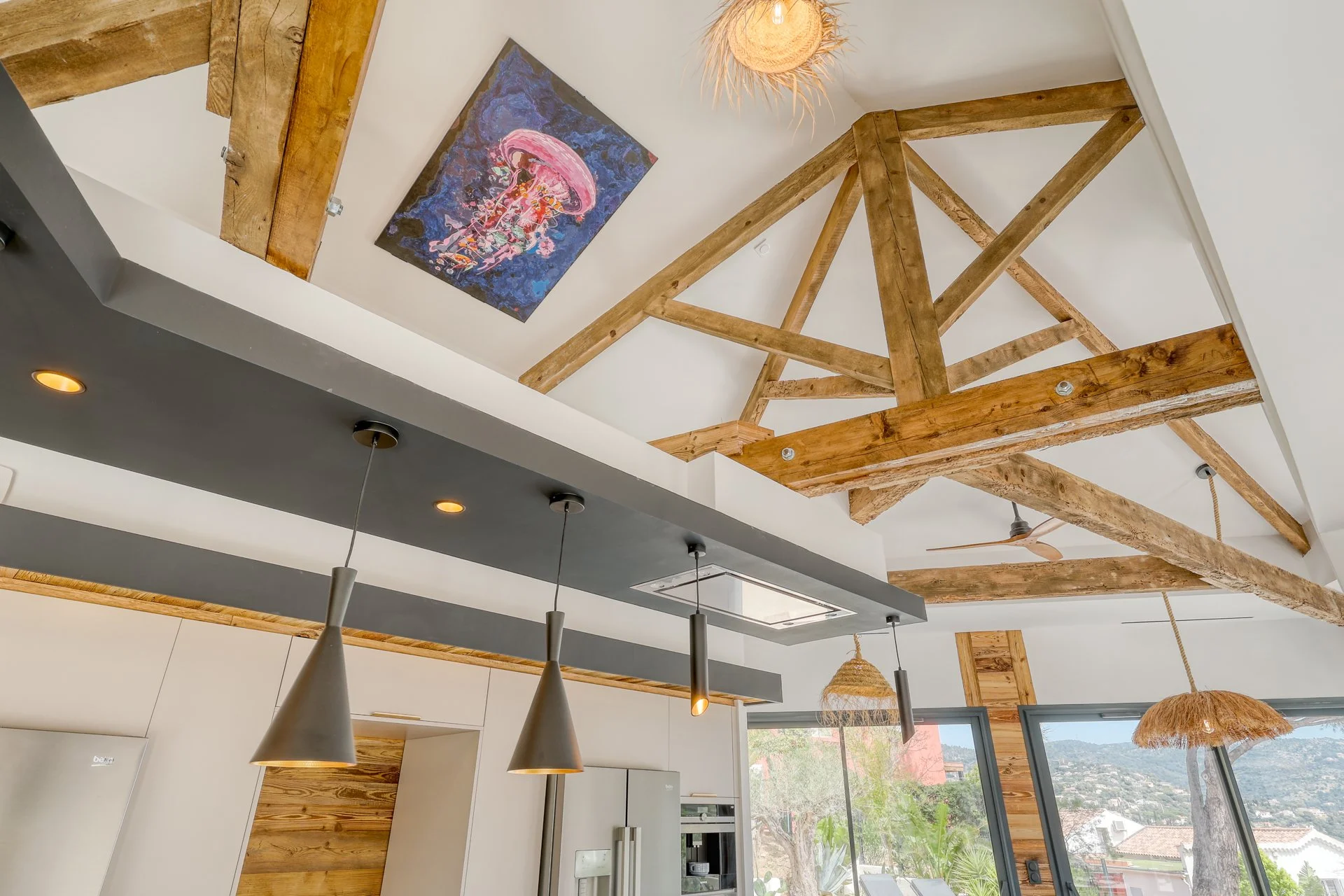 Intérieur moderne d'une maison avec plafond haut, poutres en bois apparentes, peinture murale représentant une méduse, fenêtres panoramiques et luminaires suspendus.