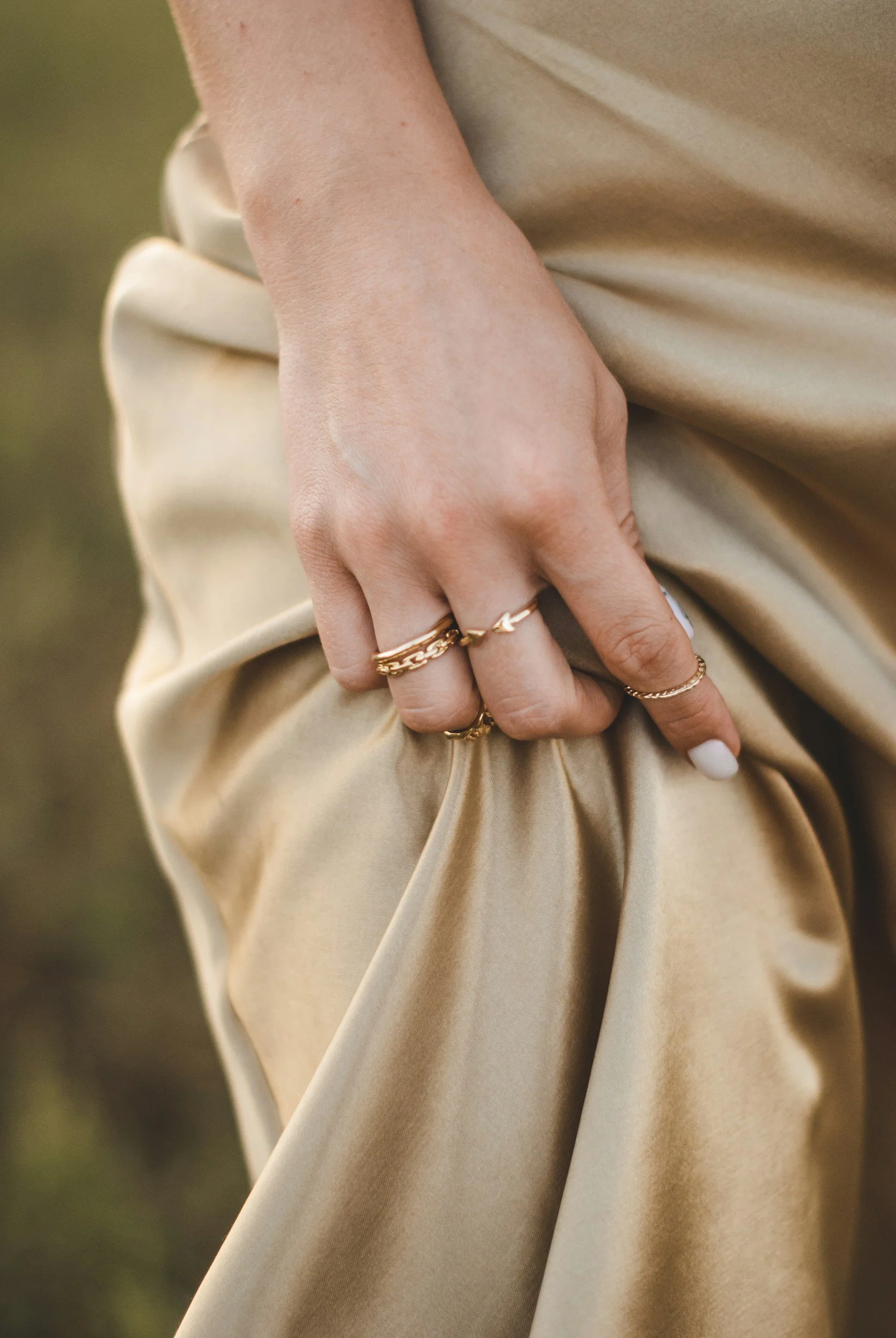 Nahaufnahme einer Hand mit mehreren goldenen Ringen, die an einem beige Stoffkleid befestigt ist, mit einem unscharfen grünen Hintergrund.