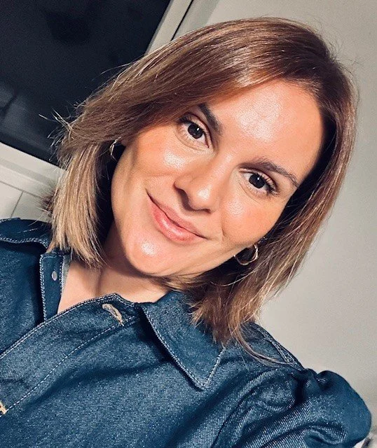 Nahaufnahme einer lachenden jungen Frau mit mittellangen, braunen Haaren, in einem dunkelblauen Denim-Hemd, vor einem Fenster in einem helle Raum.