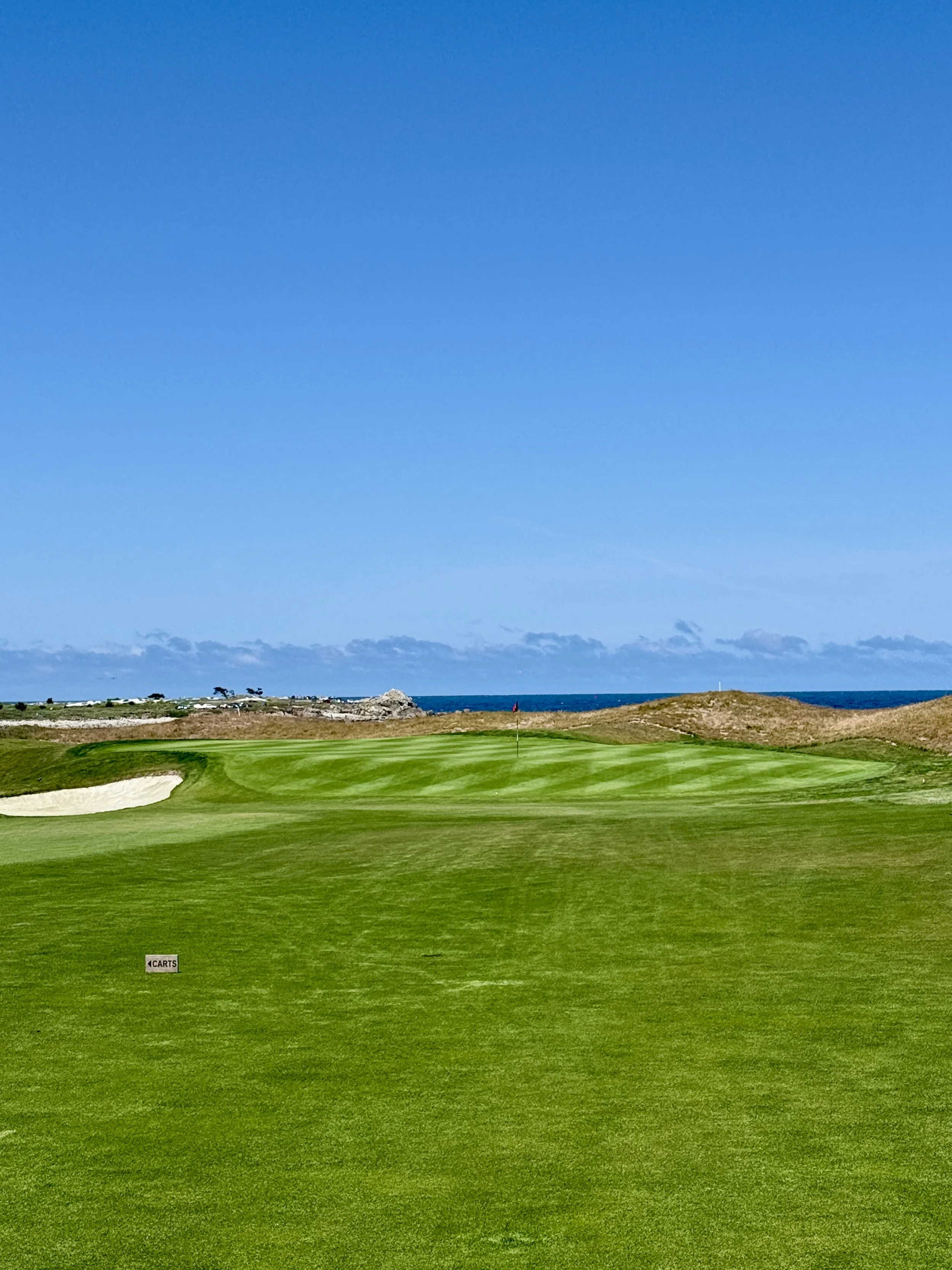 Een golfterrein met groene fairways, sandbunkers en een vlag op de green, uitzicht op de oceaan en een blauwe hemel met enkele wolken in de achtergrond.