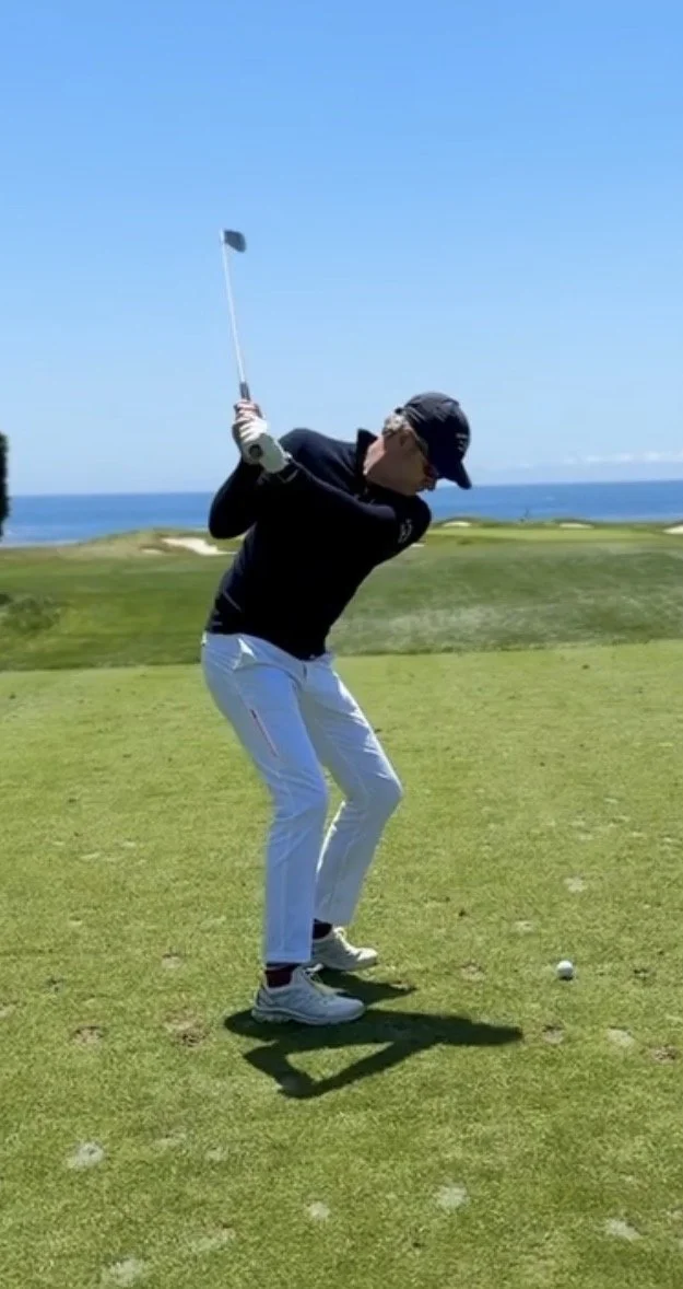 Een persoon die golf speelt op een grasveld met uitzicht op de zee, klaar om een golfslag te slaan.