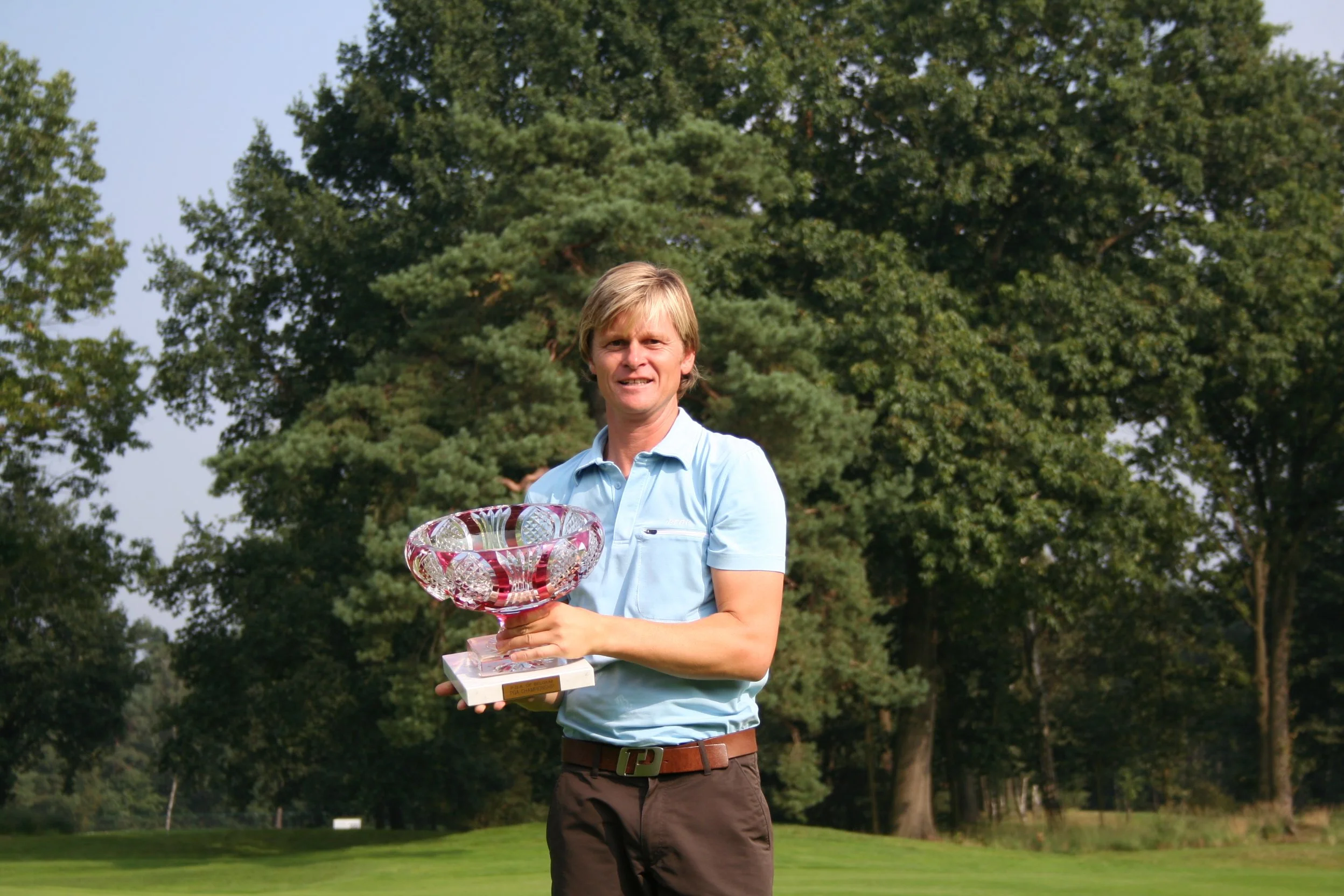 Man staat op een grasveld met bomen op de achtergrond, hij houdt een grote glazen trofee vast en kijkt tevreden.
