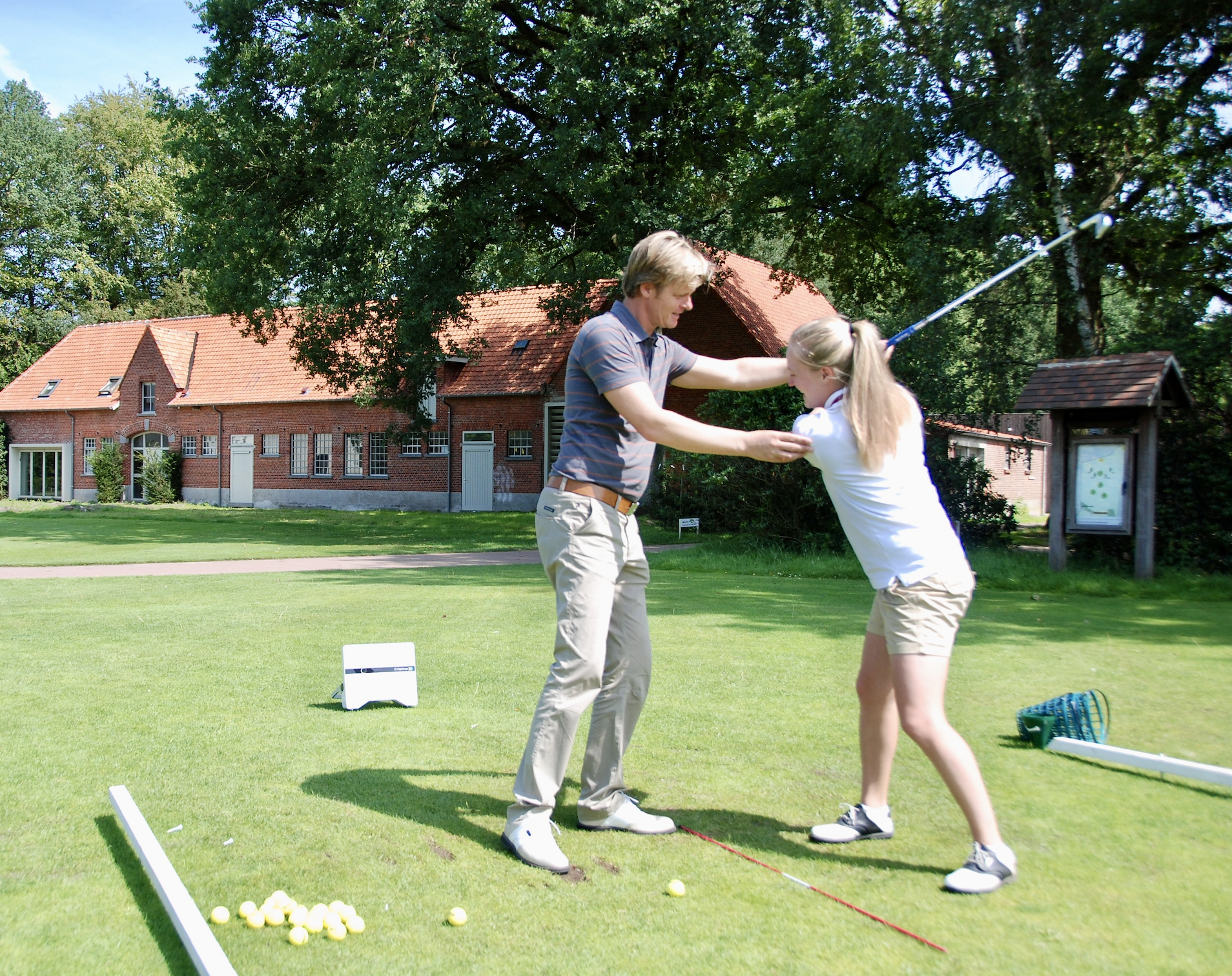 Een man en een meisje oefenen golf op een grasveld met een huis en grote bomen op de achtergrond. De man helpt het meisje met haar swing, terwijl ze zich voorbereidt om de bal te slaan.