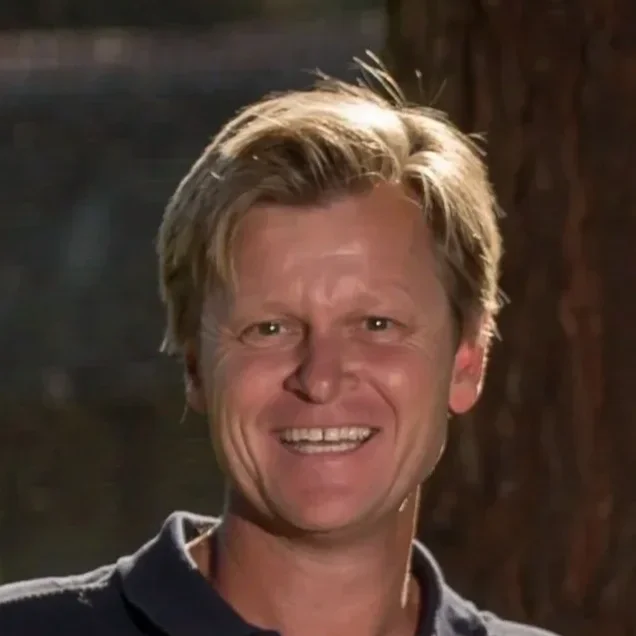 Een lachende man met blonde haar, gesitueerd in een natuurlijke omgeving.