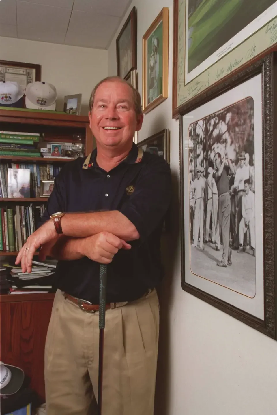 Een lachende man die in een kamer staat met zijn armen over elkaar, omringd door boeken en muurdecoraties, met een golfclub in zijn hand.