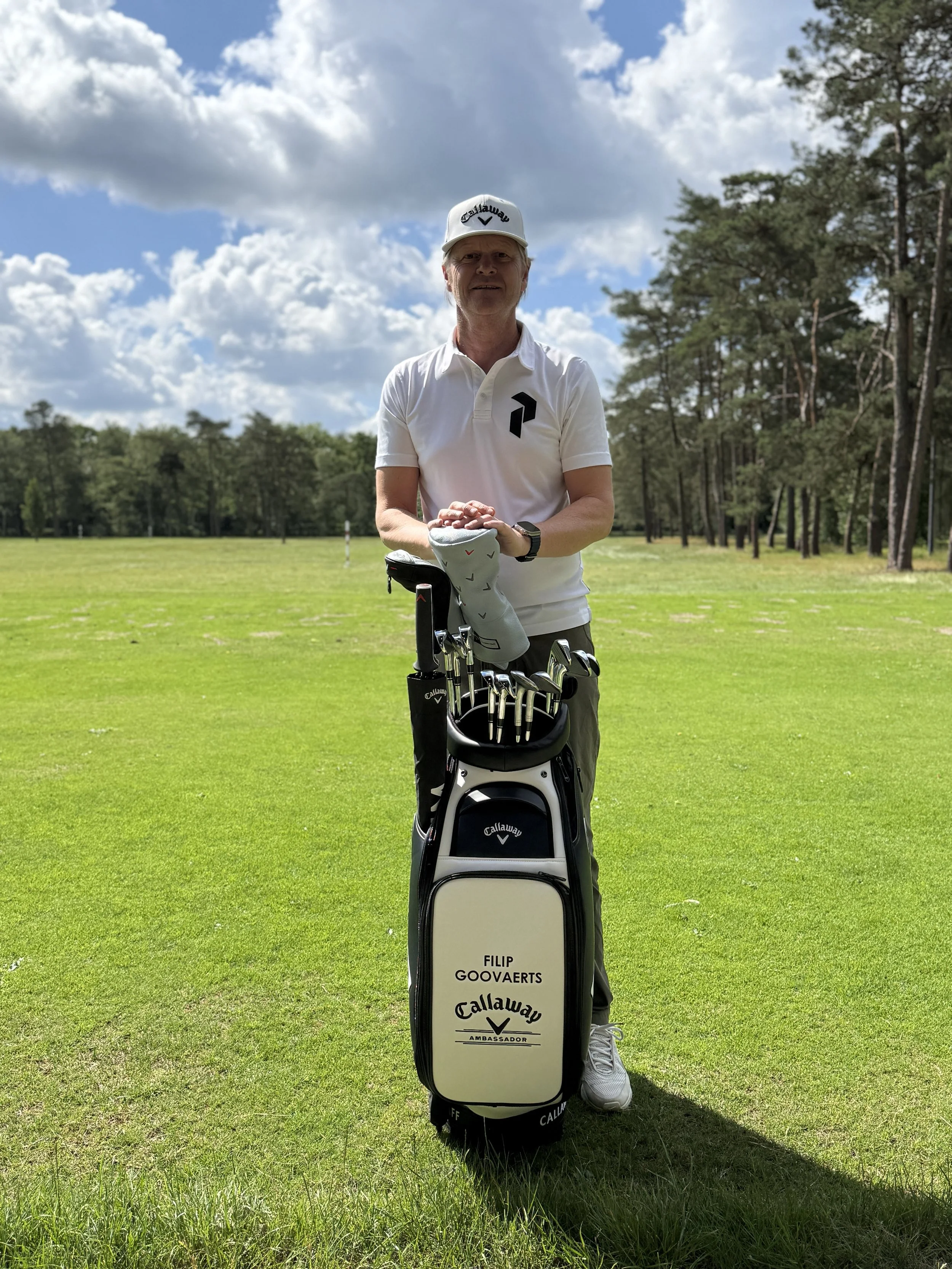Man staat op een golfbaan met een golfbeker en golfclubs, draagt een wit golfshirt en een witte pet, en heeft een soloreis met een bosrijke achtergrond onder een bewolkte blauwe hemel.