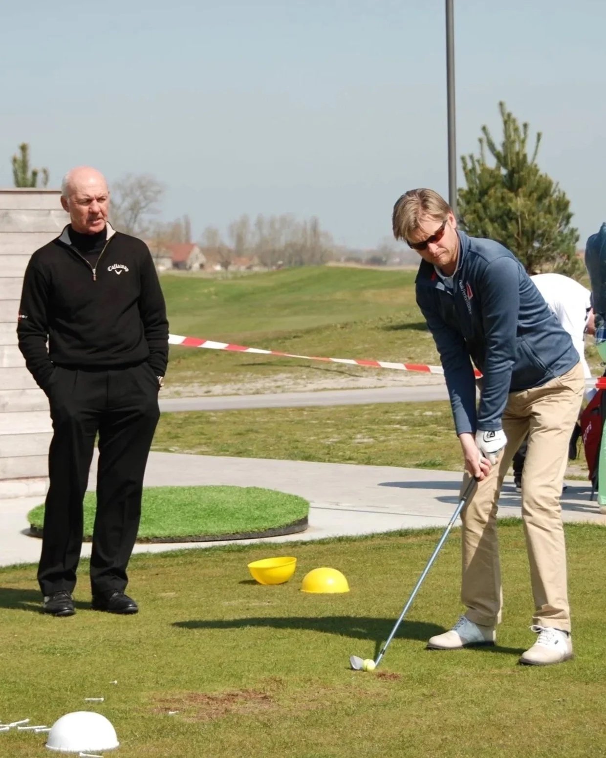 Man die golf speelt op een golfbaan, met twee gele markeringen en mensen die toekijken, en een man die zich voorbereidt om te slaan.
