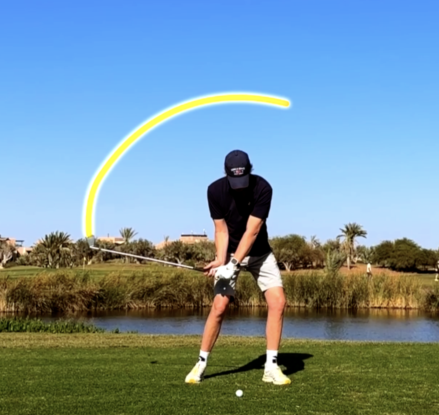 Man die een golfmove uitvoert op een golfbaan met blauwe lucht en palmbomen op de achtergrond.