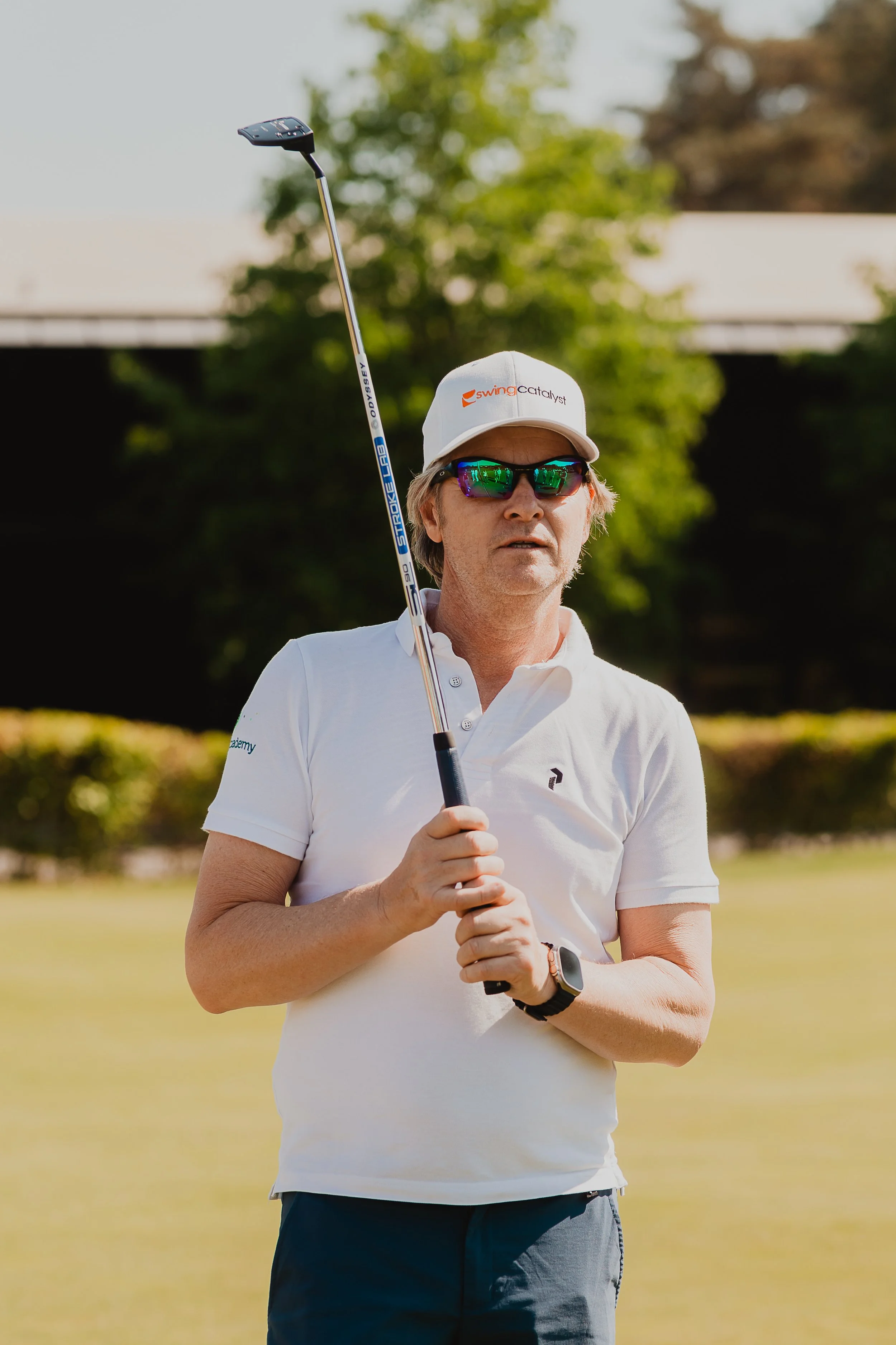 Een man met een witte polo en een witte pet die op een grasveld staat, omgeven door bomen, terwijl hij een golfclub vasthoudt.