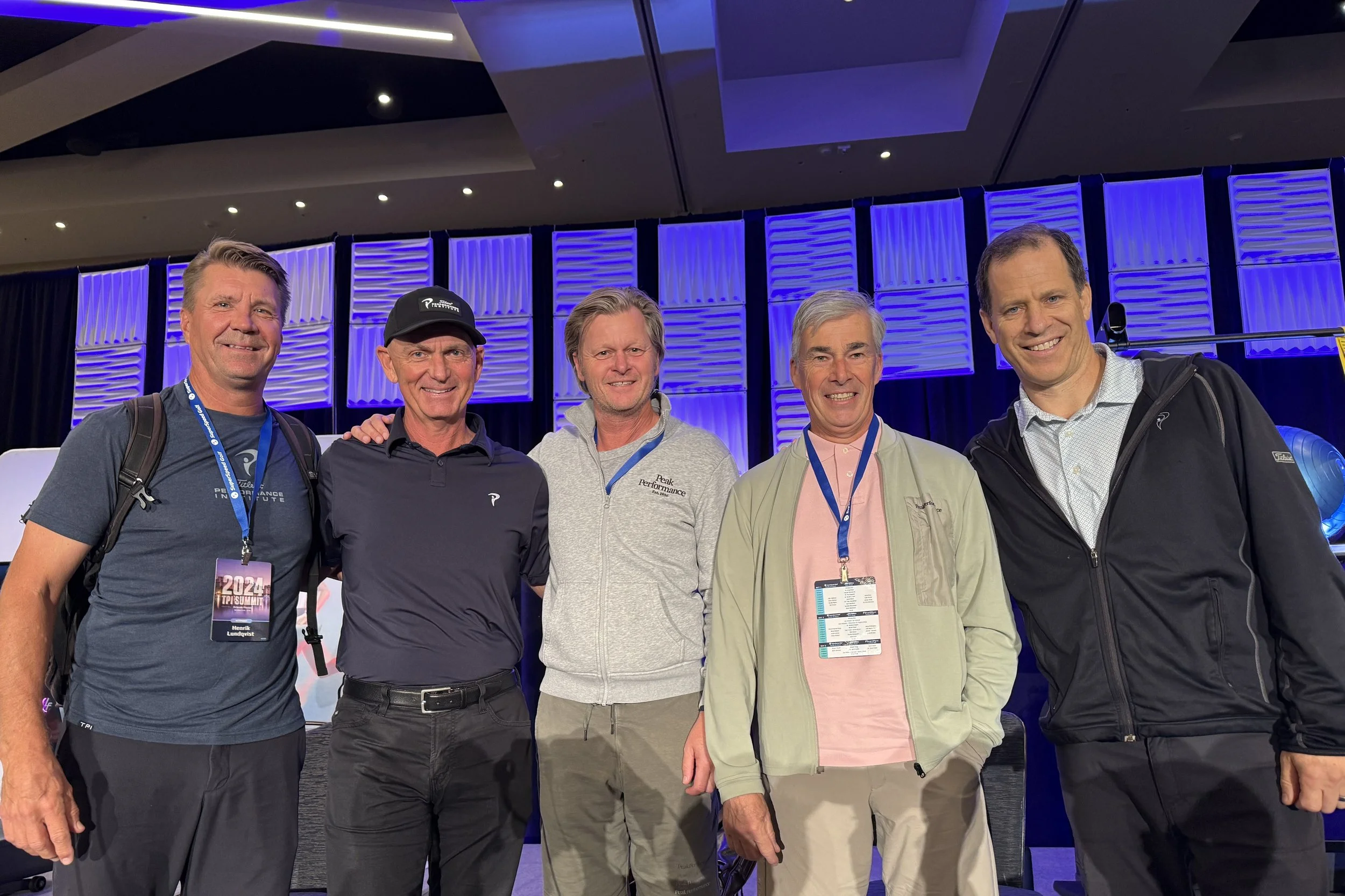 Vijf mannen staan samen op een conferentie of evenement, gekleed in casual en zakelijke kleding, met blauwe en paarse achtergrondverlichting.