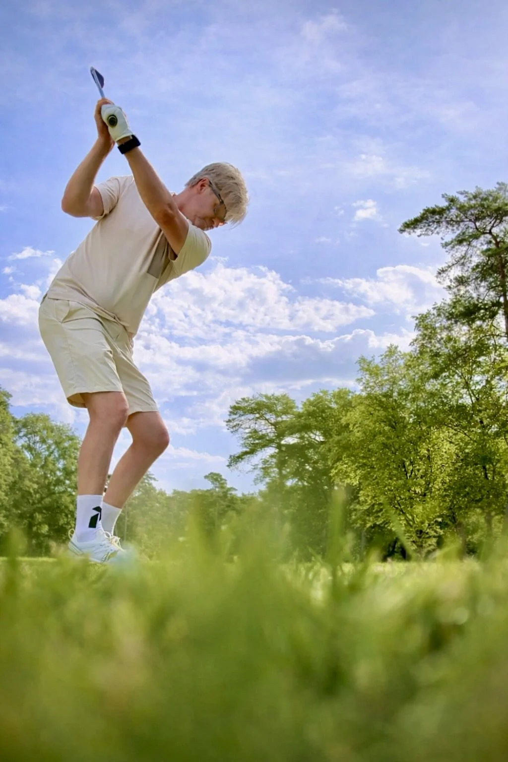 Man die een golf slaat op een golfbaan met groene bomen en een blauwe lucht op de achtergrond.
