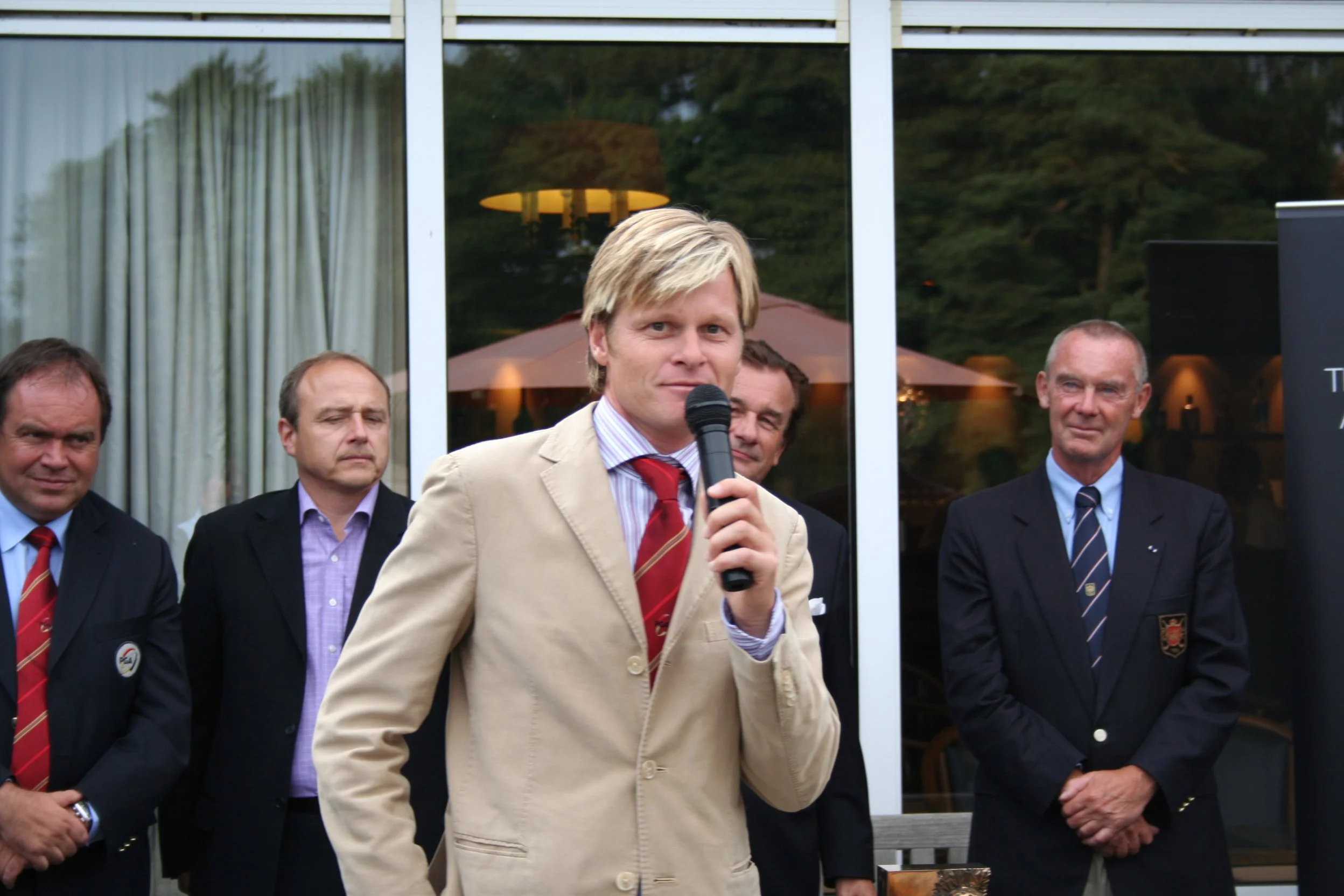 Man in beige suit enrodeert met microfoon, omringd door mannen in formele kleding, buiten bij een gebouw met grote ramen.