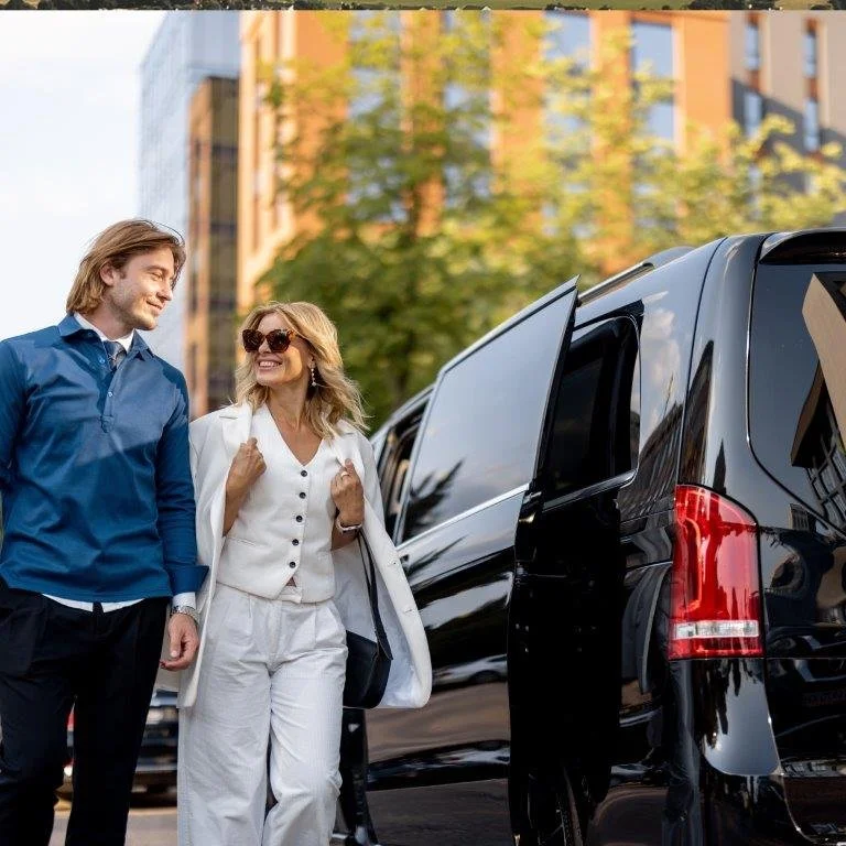 A man and woman walking on a city street, smiling and looking at each other, with buildings and trees in the background. The woman is wearing sunglasses and a white outfit, while the man is dressed in a blue shirt.