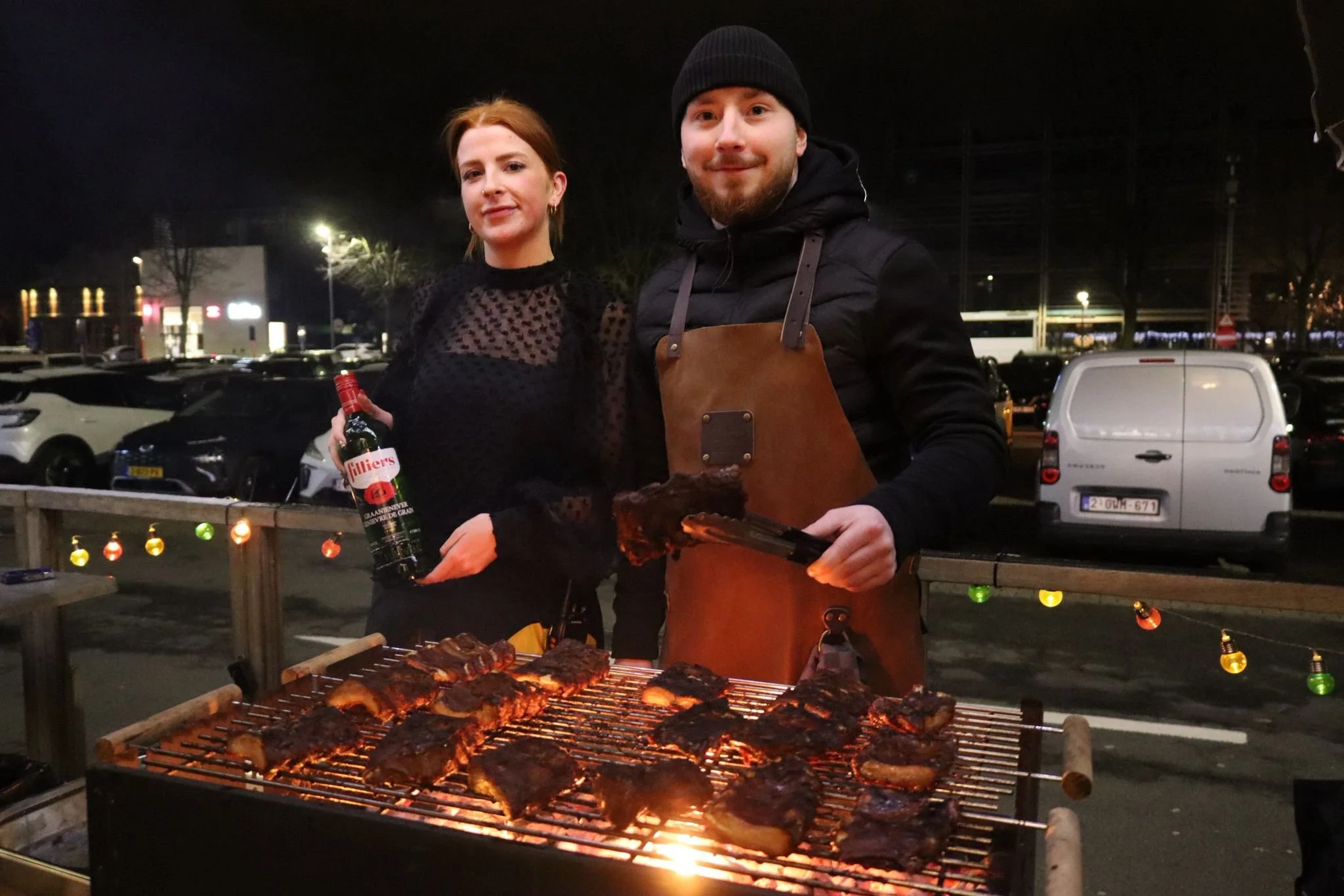 A man and woman standing behind a grill with meat, celebrating outdoors at night in a parking lot decorated with colorful string lights, with cars parked in the background.