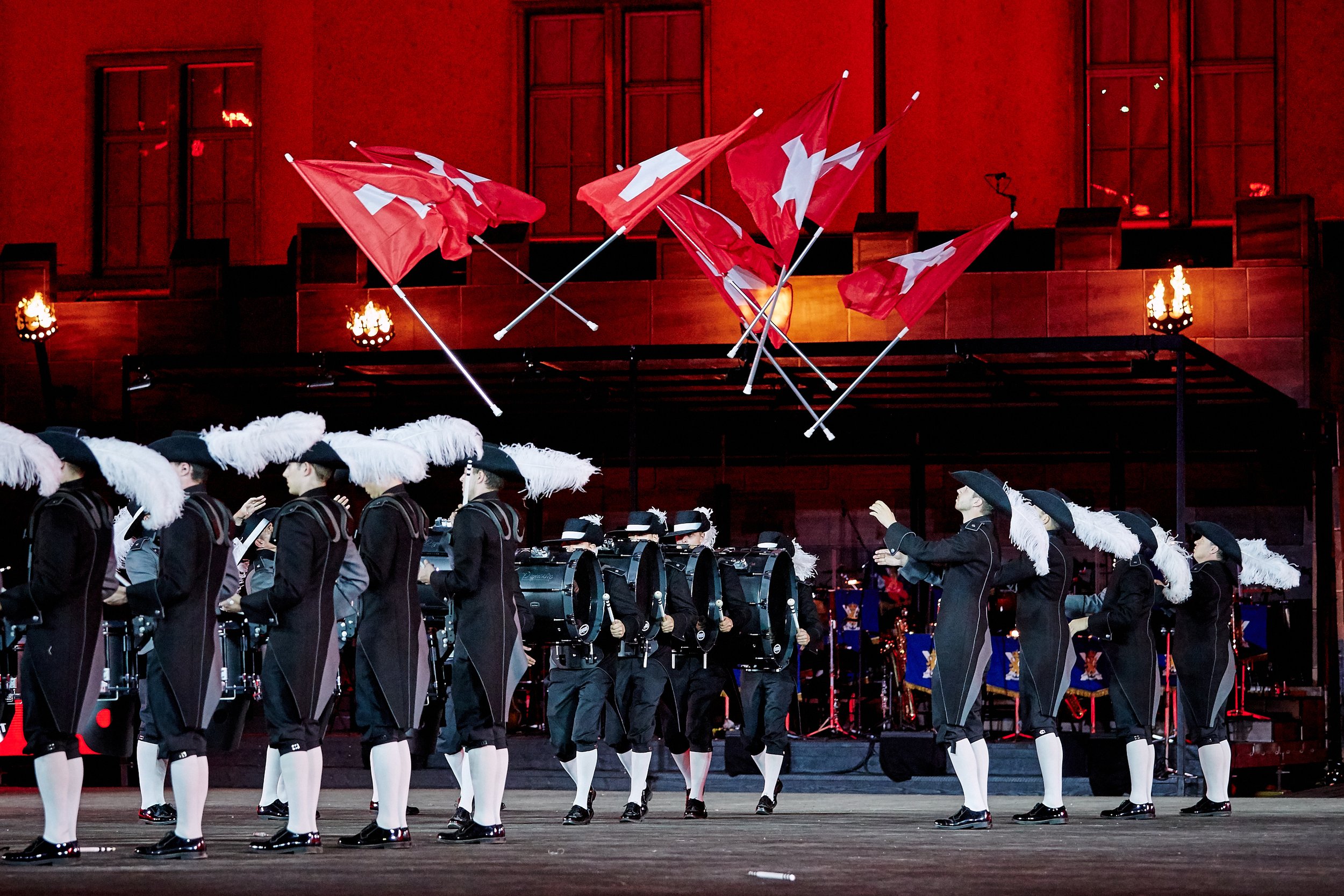 Eine Gruppe von Musikern oder Marschkapellenmitgliedern in historischen Uniformen mit Federhauben, die auf einer Bühne auftreten, mit mehreren roten Schweizer Flaggen im Hintergrund.