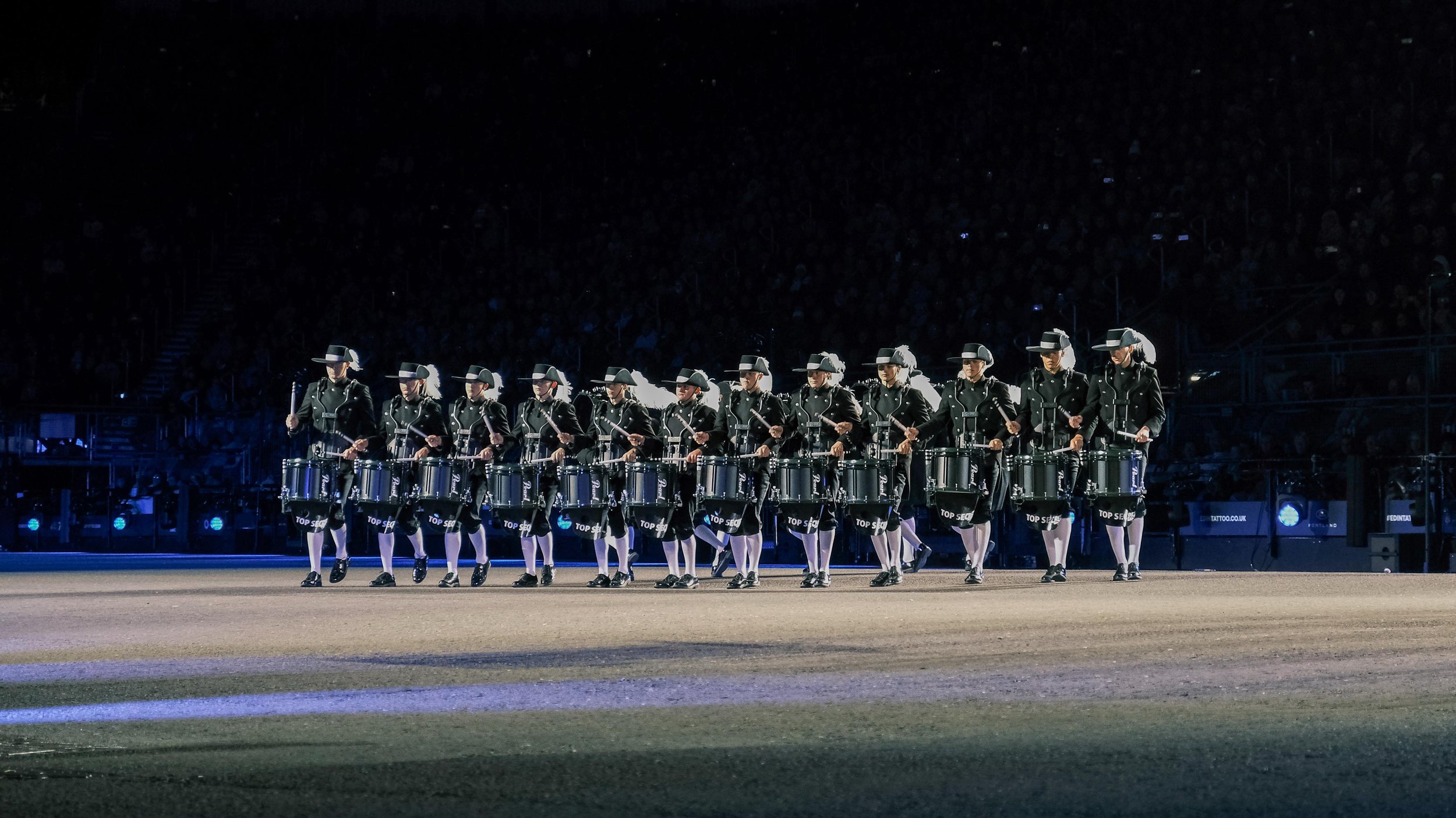 Eine Gruppe von Trommlern in schwarzen Uniformen und Hüten, die einen Marschtrommel-Aufmarsch in einem dunklen Stadion durchführen.