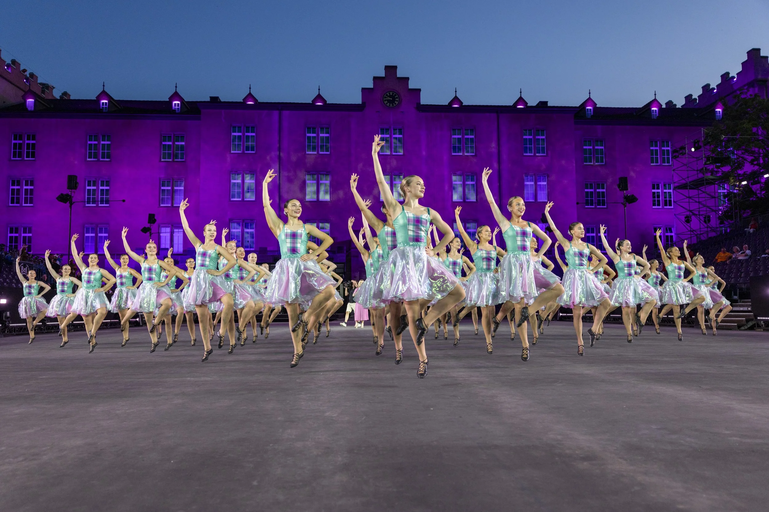 Tänzerinnen in bunten Kostümen beim Tanz auf einer Bühne vor einem pink beleuchteten Gebäude während einer Aufführung bei Nacht.