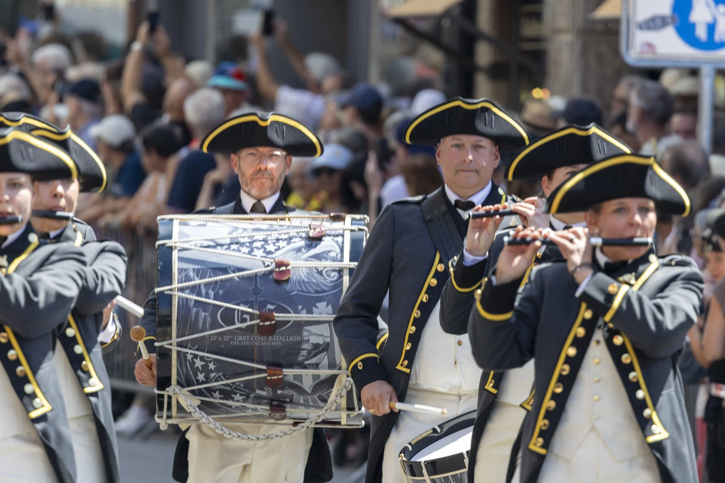 Musiker in historischer Kleidung bei einer Parade, spielen Flöte und Trommel, im Hintergrund eine Menschenmenge und eine Straße.