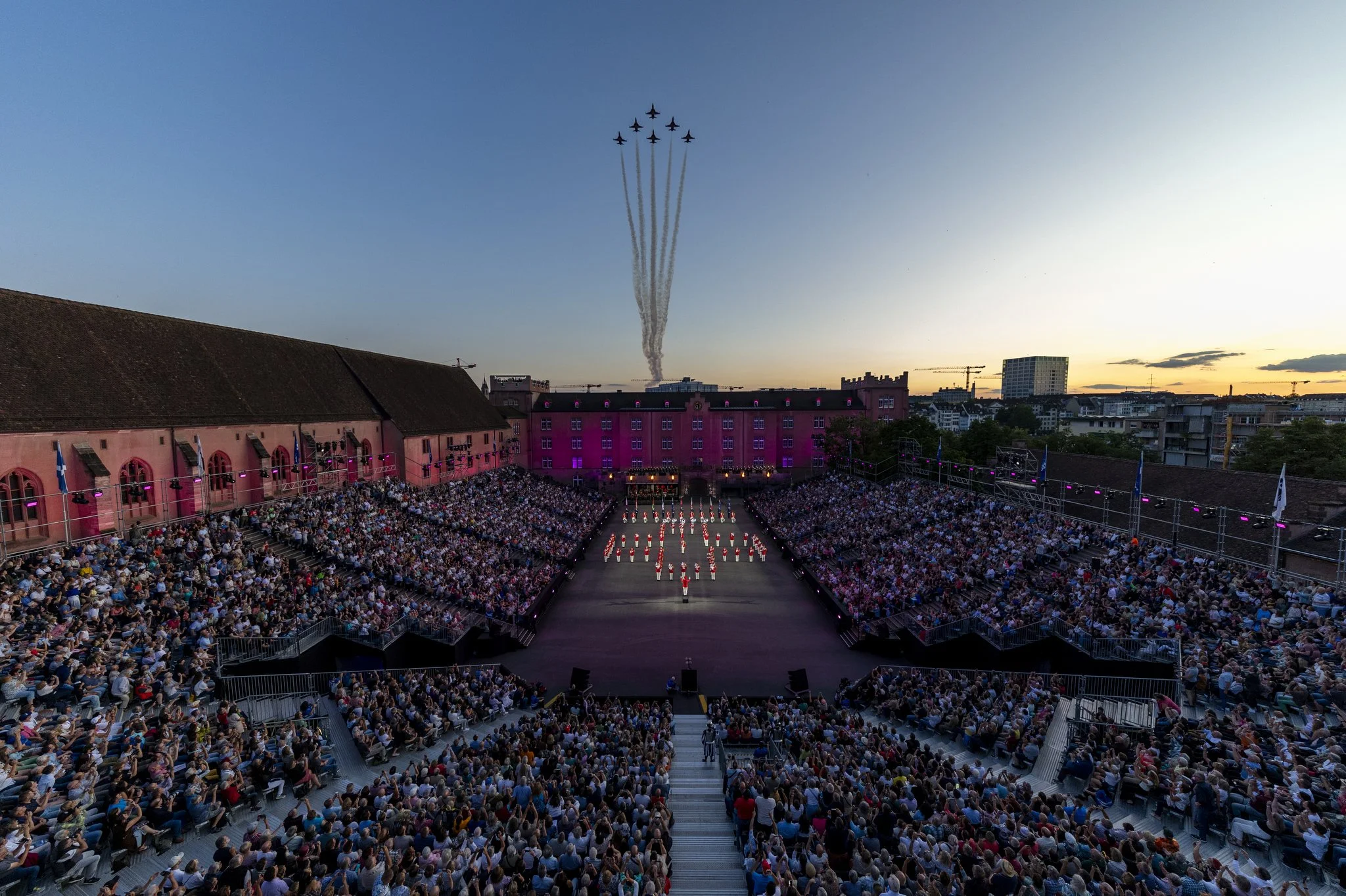 Aerial view of a large outdoor event with a lit-up stage, surrounded by a seated audience, pink building in the background, and fighter jets flying in formation across the sunset sky.
