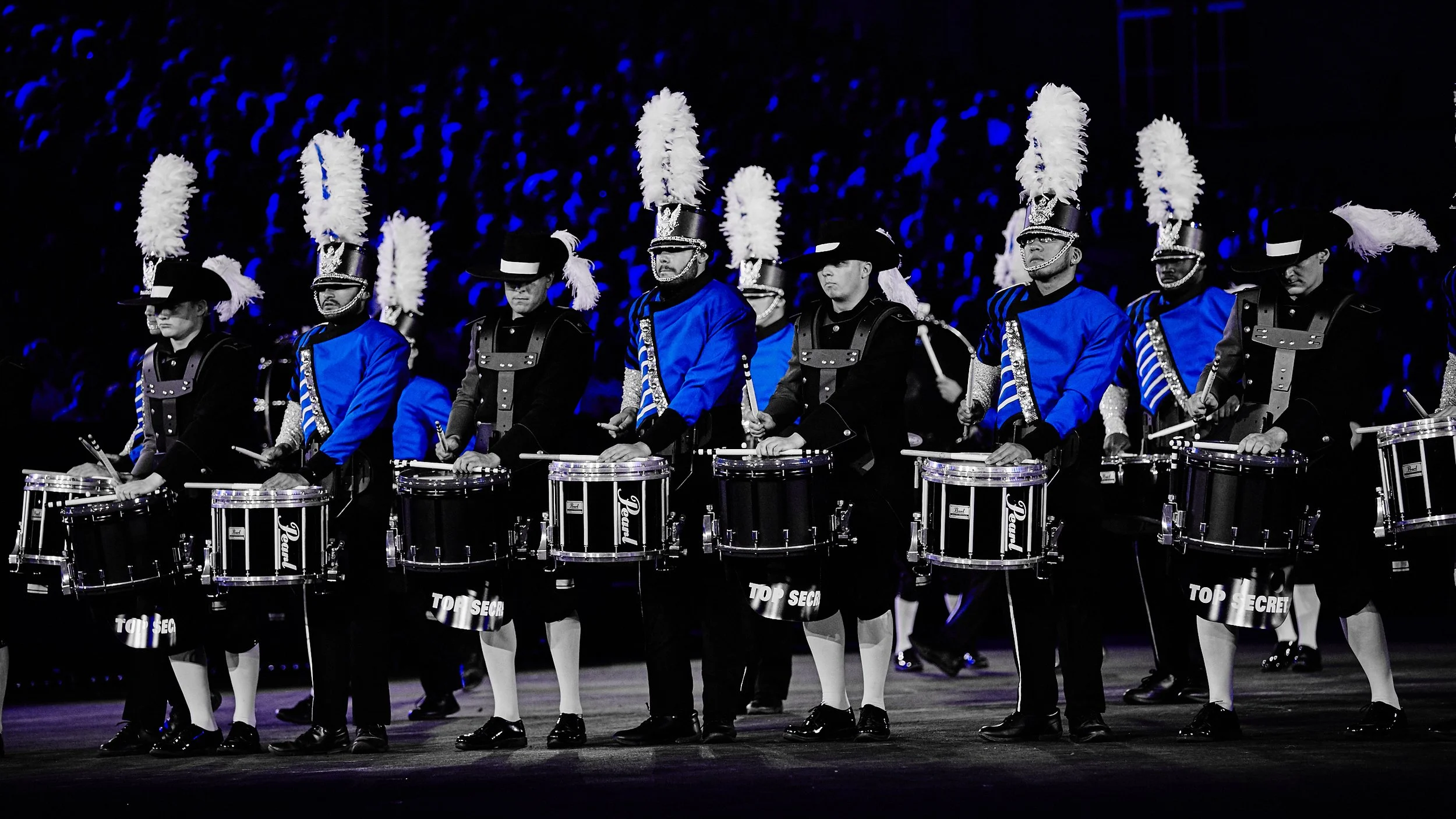 Musiker in historischen Kostümen mit schwarzen und blauen Anzügen, die Trommeln spielen, in einer Dunkelheit bei einem feierlichen Ereignis, mit bläulicher Beleuchtung im Hintergrund.